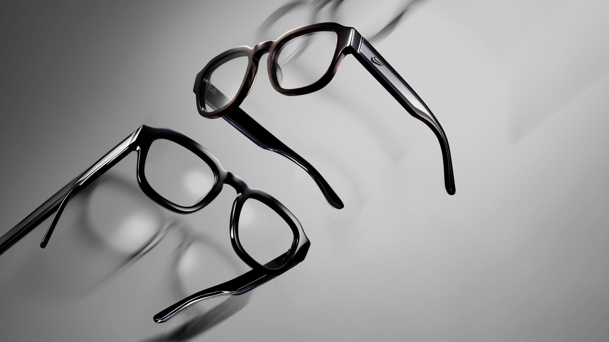 Two pairs of eyeglasses, one pair with tortoiseshell frames and the other with black frames, placed on a reflective surface with a gray background.