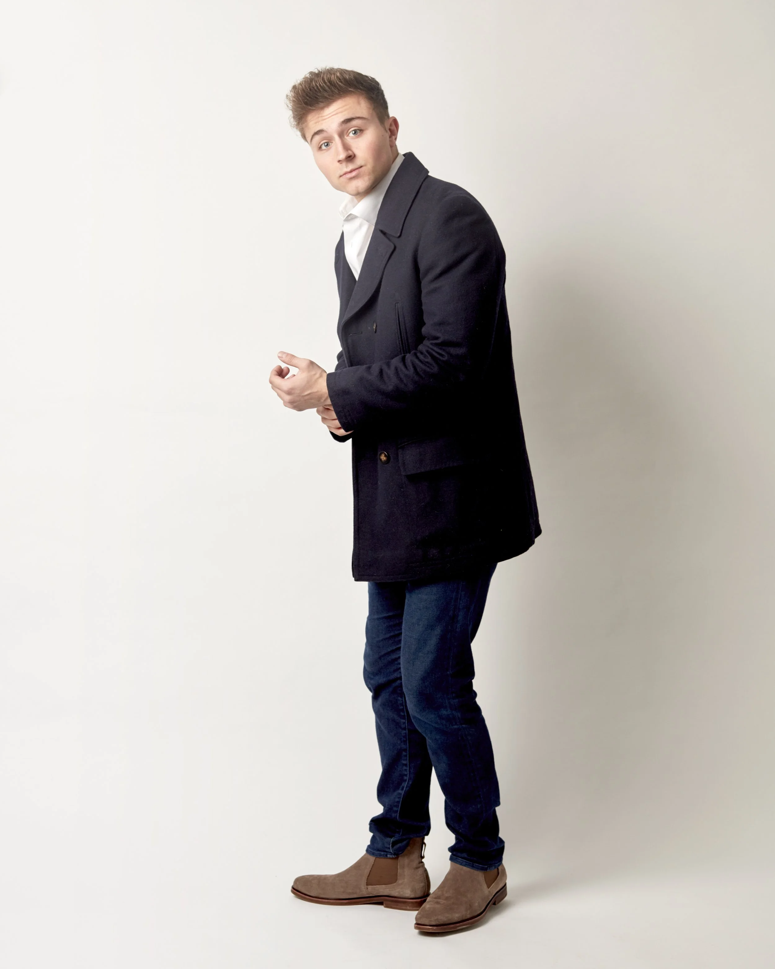 Young man with light brown hair dressed in a dark coat, white shirt, jeans, and tan boots, standing against a plain white background, looking at the camera.