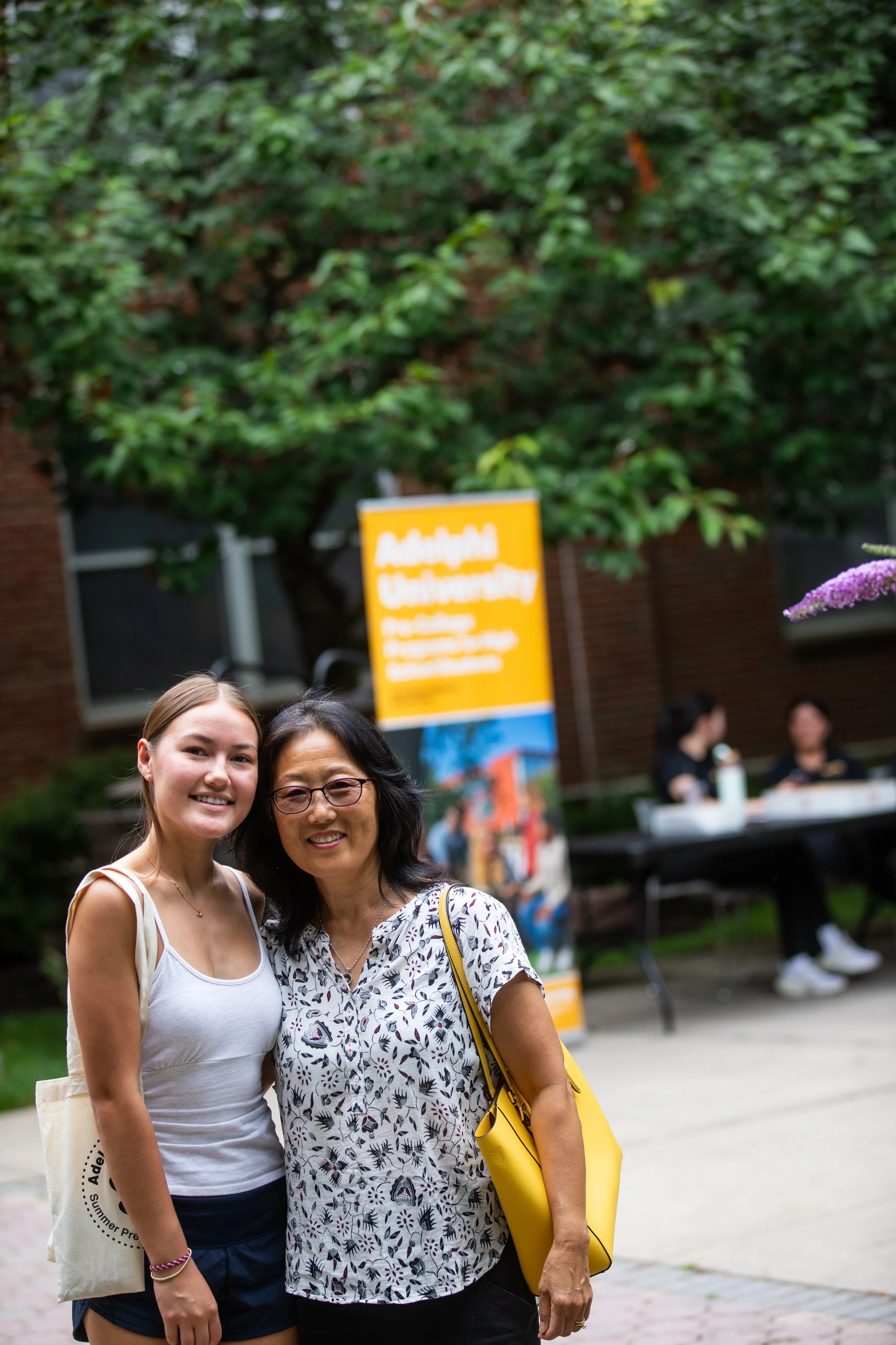 Two women standing together outdoors at Adams University, smiling at the camera. One woman is young with light skin and blonde hair, wearing a white tank top and navy shorts. The other woman is older with darker skin and black hair, wearing glasses, 