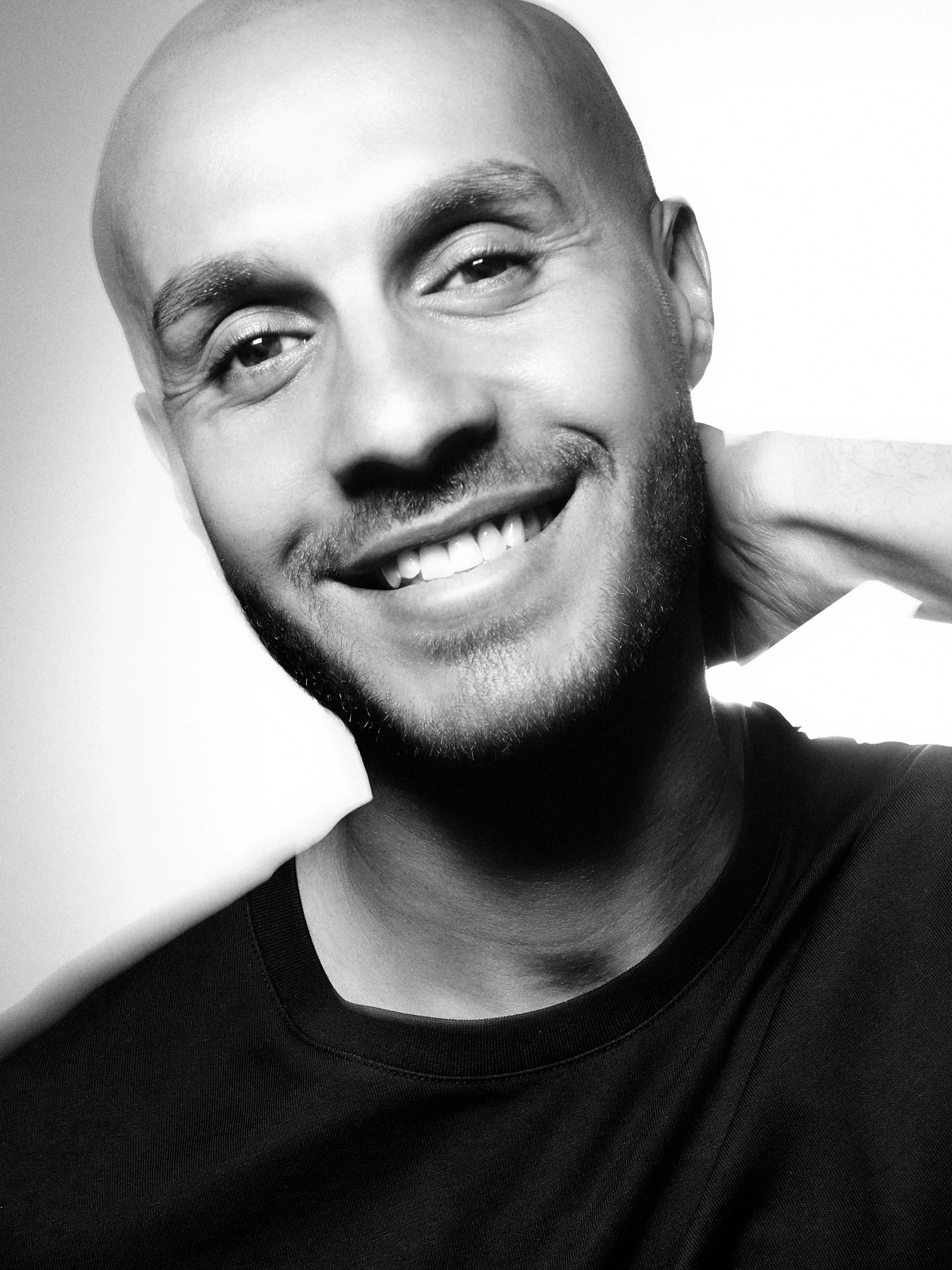 A smiling man with a shaved head and a beard, resting his hand on the side of his face, wearing a dark shirt.