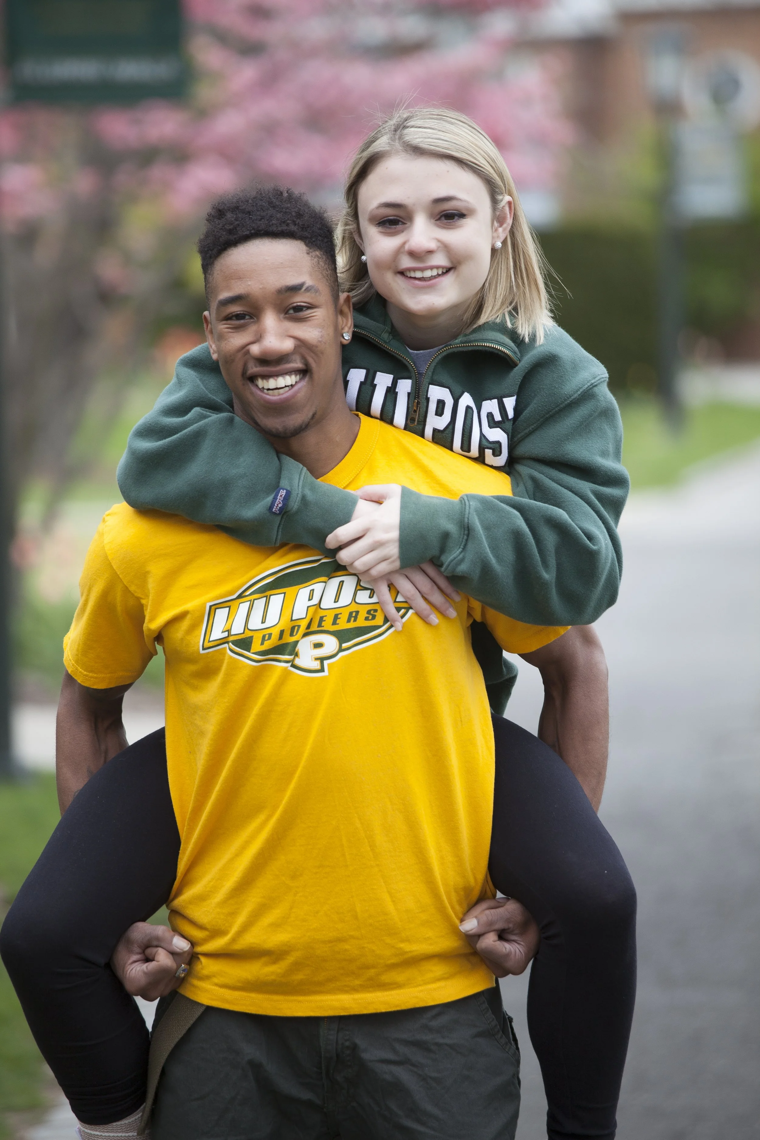 A young man in a yellow Purdue Boilermakers T-shirt giving a piggyback ride to a young woman wearing a green LU Post sweatshirt, both smiling, outdoors in a park with trees and pink blossoms in the background.