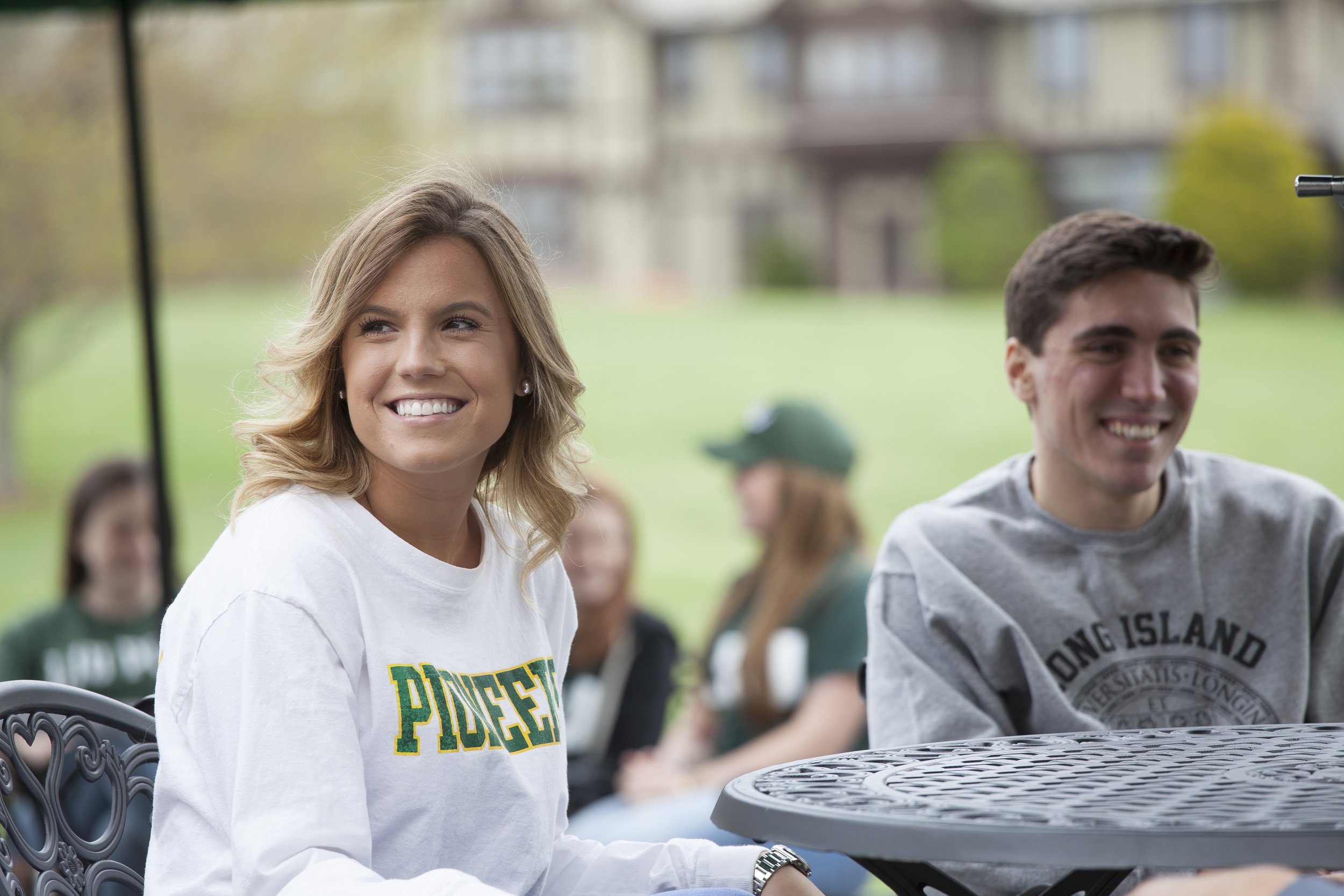 Smiling young woman with blonde hair wearing a white shirt with green and yellow text sitting outdoors with a group of people at a table in a park.