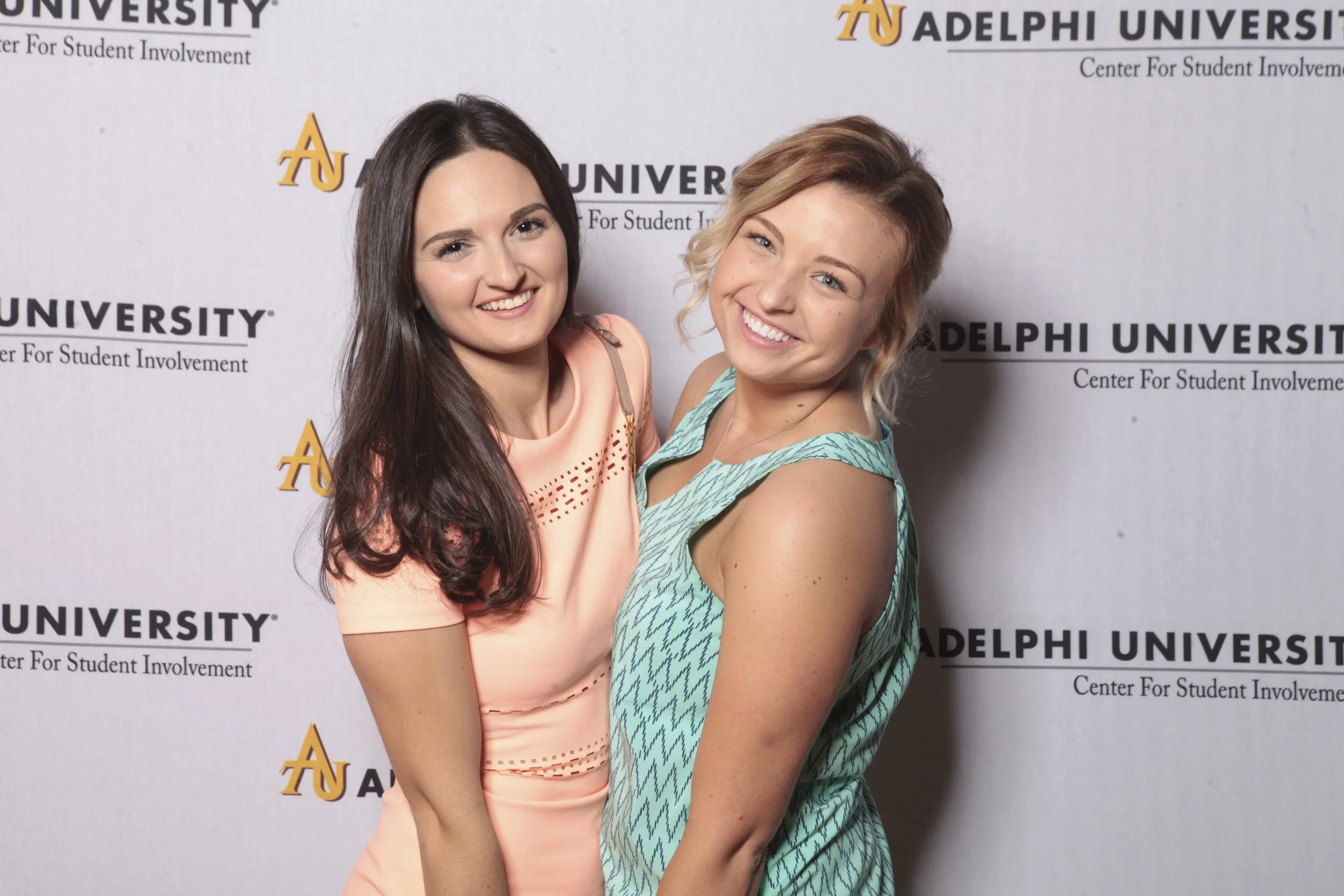 Two young women smiling and posing together at an Adelphi University event, standing in front of a backdrop with the university's logo and name.