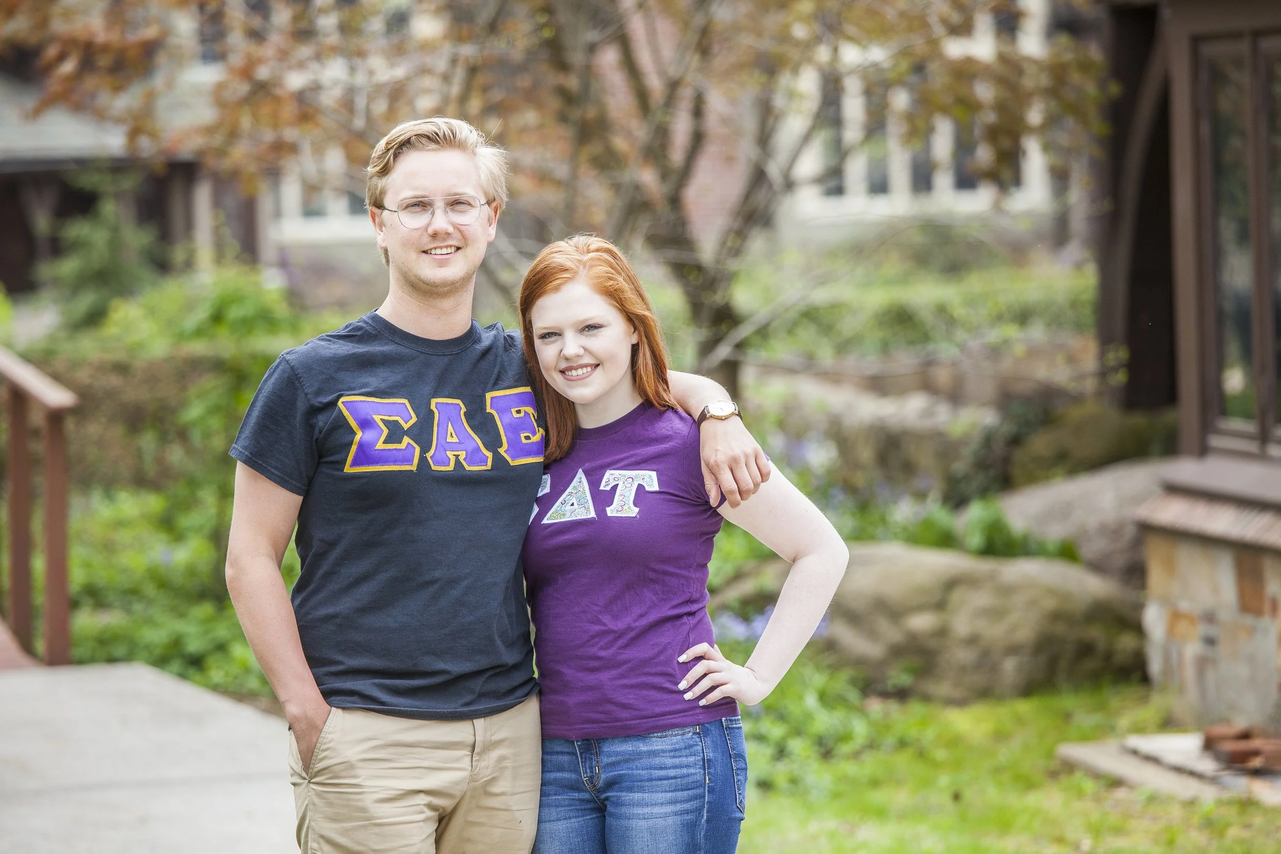 A young man and woman standing outdoors, smiling. The man has his arm around the woman, both wearing purple Greek letters on their shirts. The background features trees and a stone building.