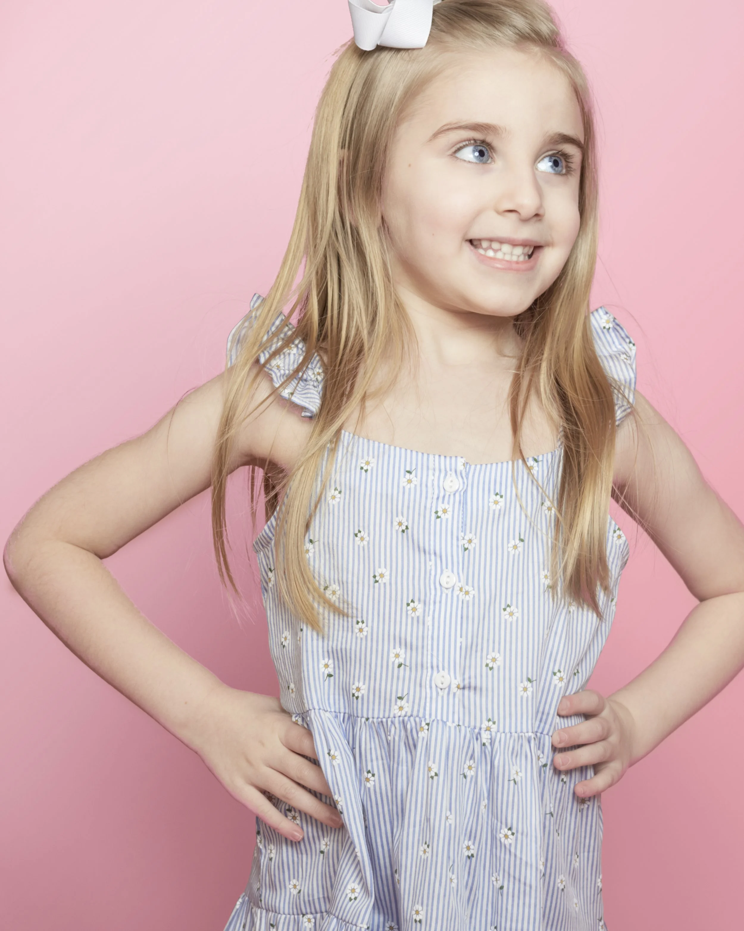 A young girl with blonde hair, blue eyes, and a white bow, standing with hands on hips, smiling, against a pink background, wearing a blue and white striped dress with small daisy patterns.