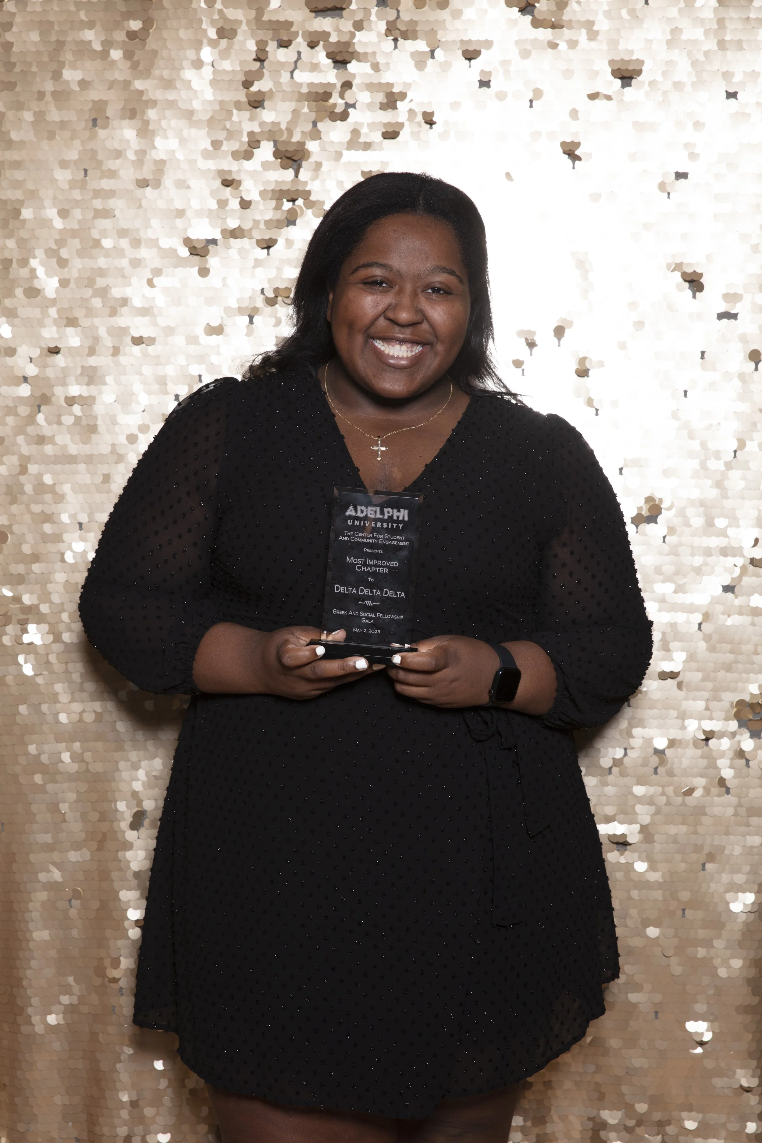 Smiling woman in black dress holding an award at Delta Delta Delta Greek and Social Fellowship Gala in front of a gold sequin backdrop.