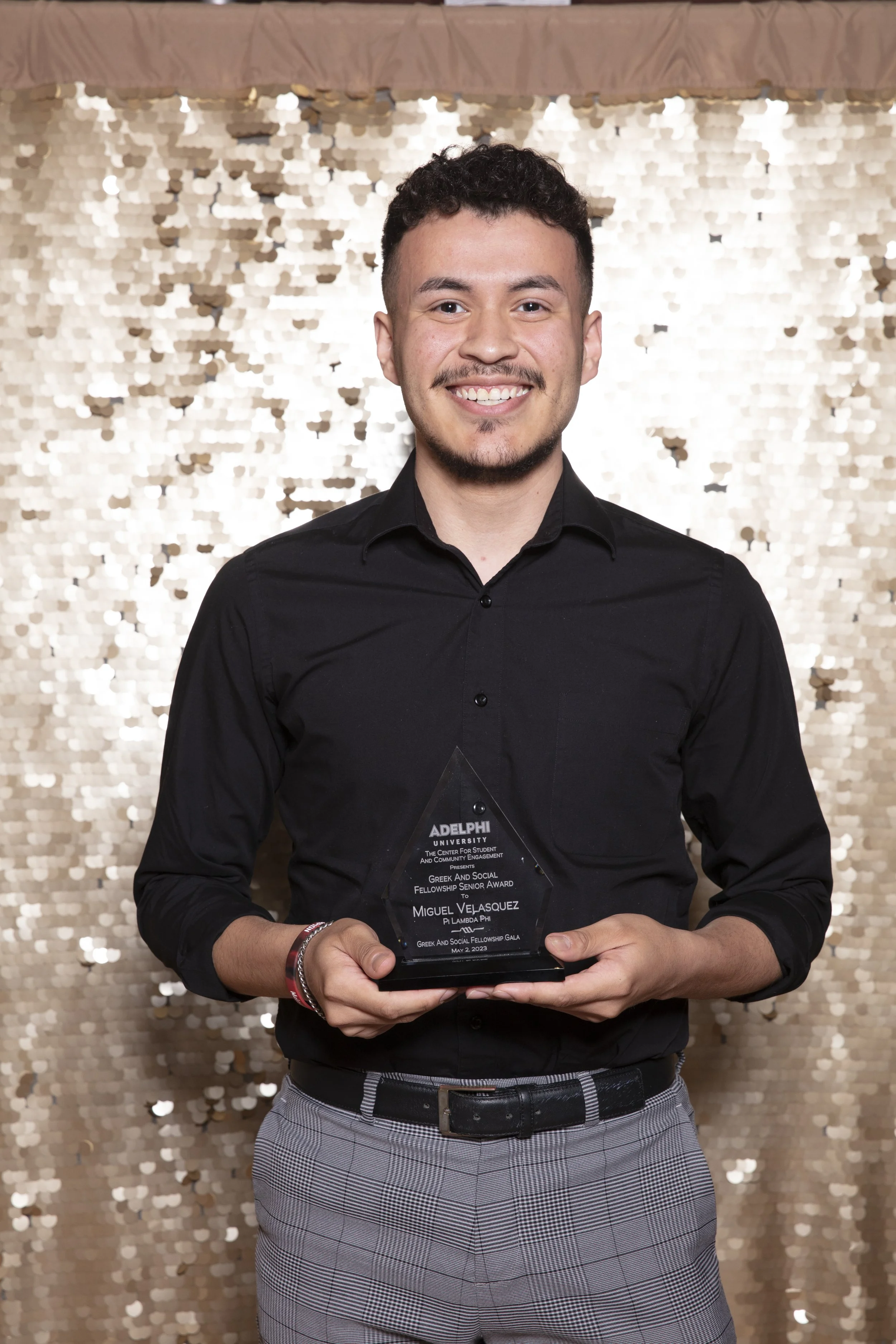 Young man holding an award at a ceremony, standing in front of a gold sequin backdrop.