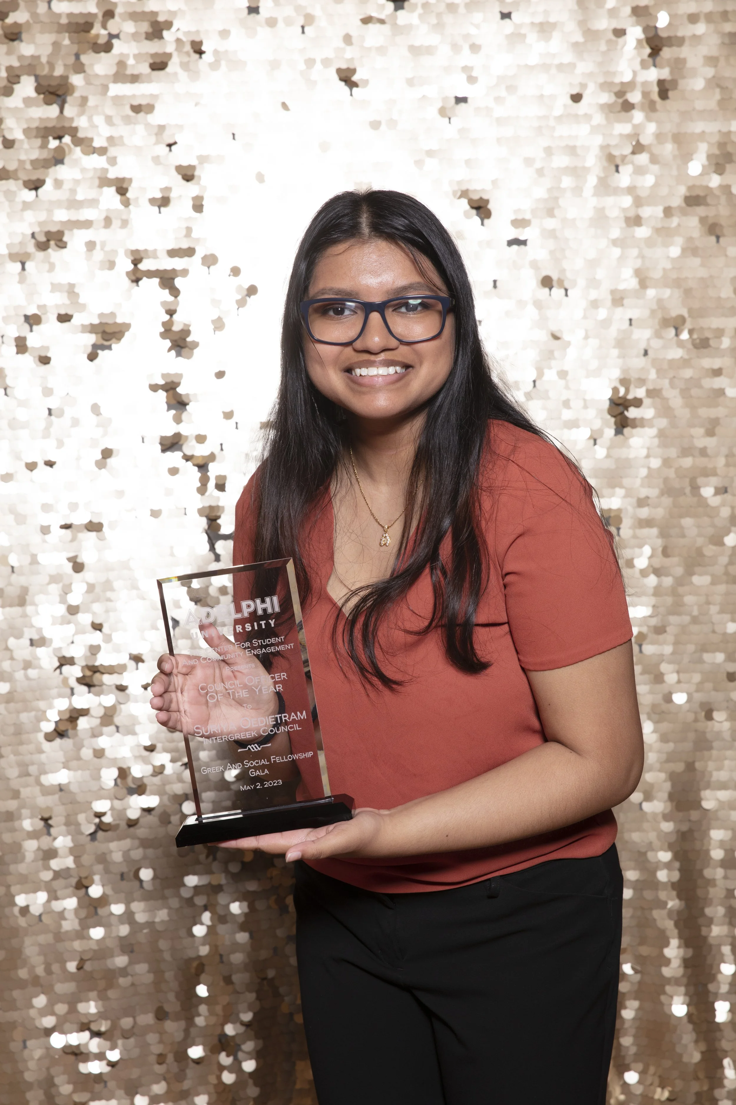 A woman with long dark hair and glasses wearing a reddish-orange shirt and black pants, smiling and holding a clear award plaque in front of a gold sequin backdrop.