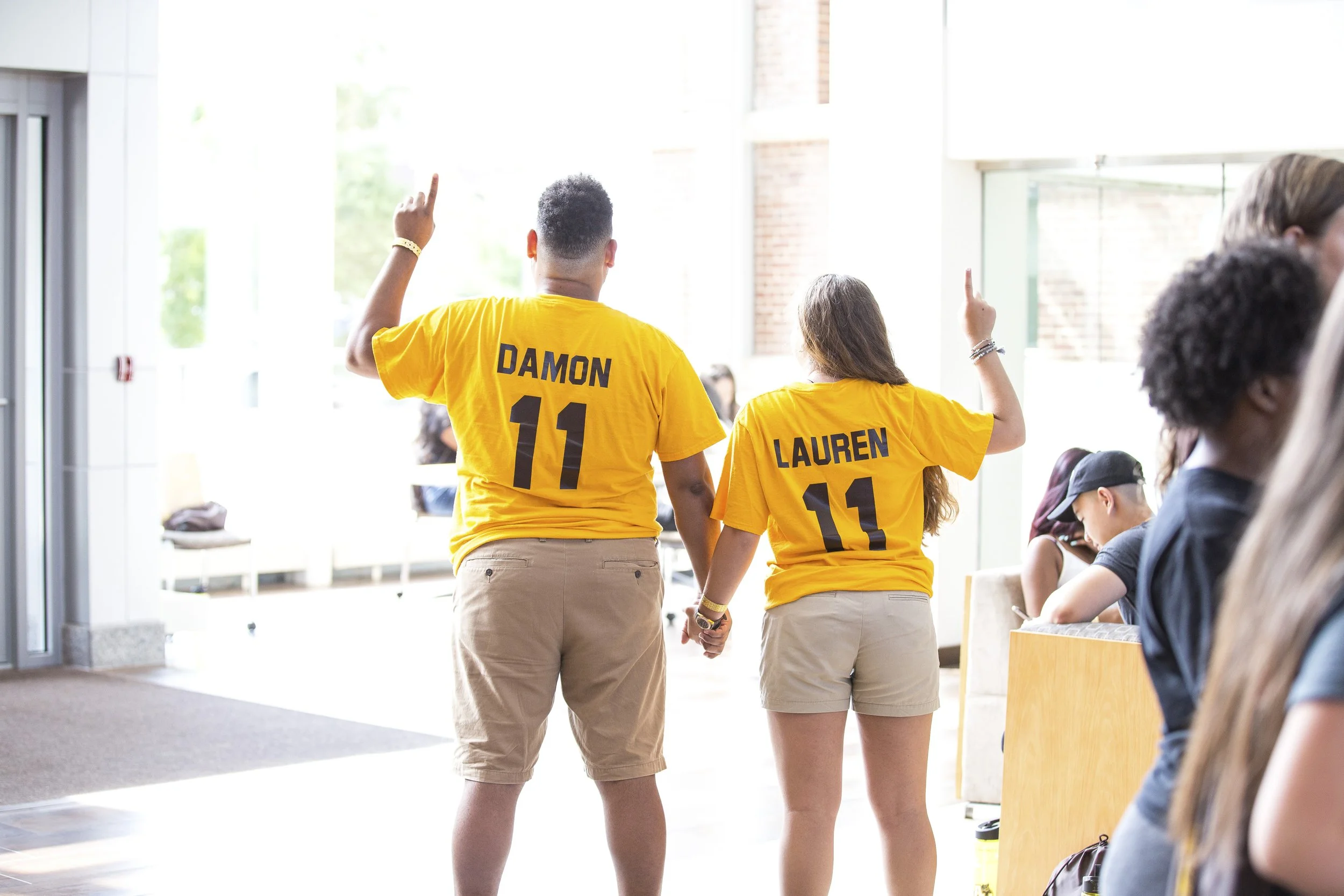 Two people, a man and a woman, wearing matching yellow shirts with the names "Damon" and "Lauren" and the number 11, holding hands, standing in a bright, modern indoor space with other people seated nearby.