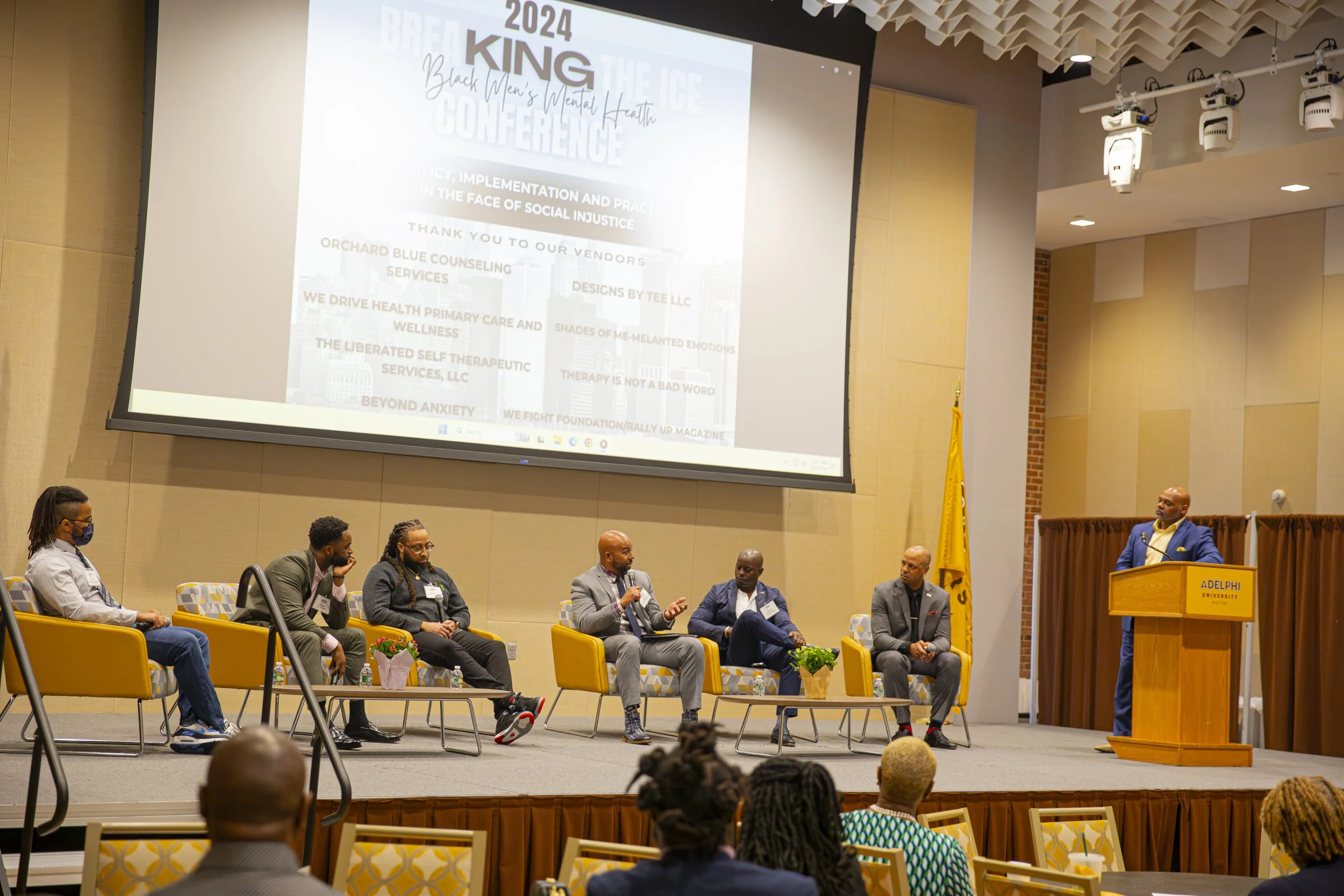 A panel discussion at a conference with six men on stage, one at a wooden podium labeled 'Adelphi University,' and five seated in chairs. A large screen behind them displays the event title '2024 KING Black Men's Mental Health Conference' with additi