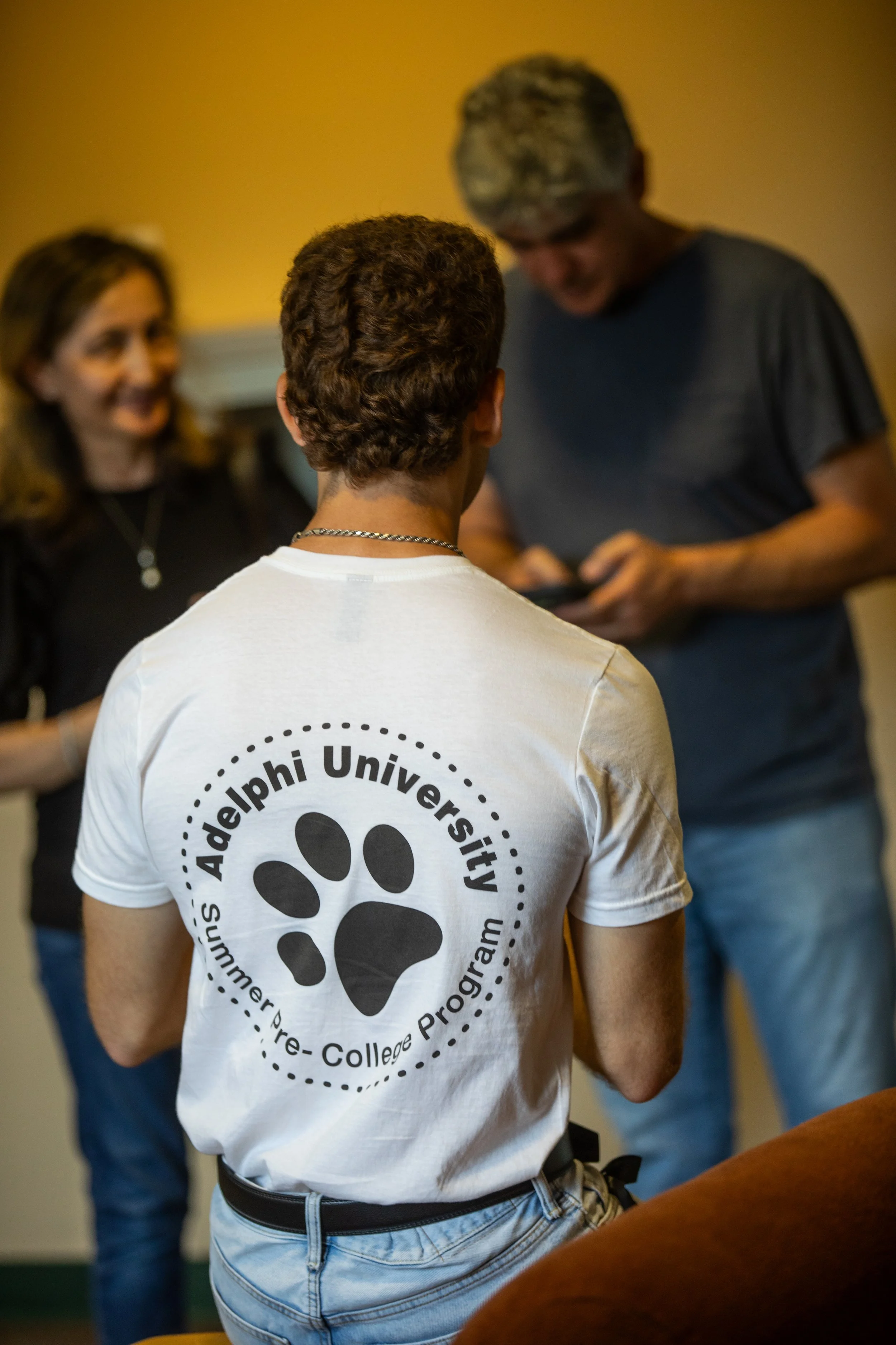 A person wearing a white t-shirt with the logo of the Summer Pre-College Program at Widener University, featuring a paw print, with two other individuals in the background talking and smiling.