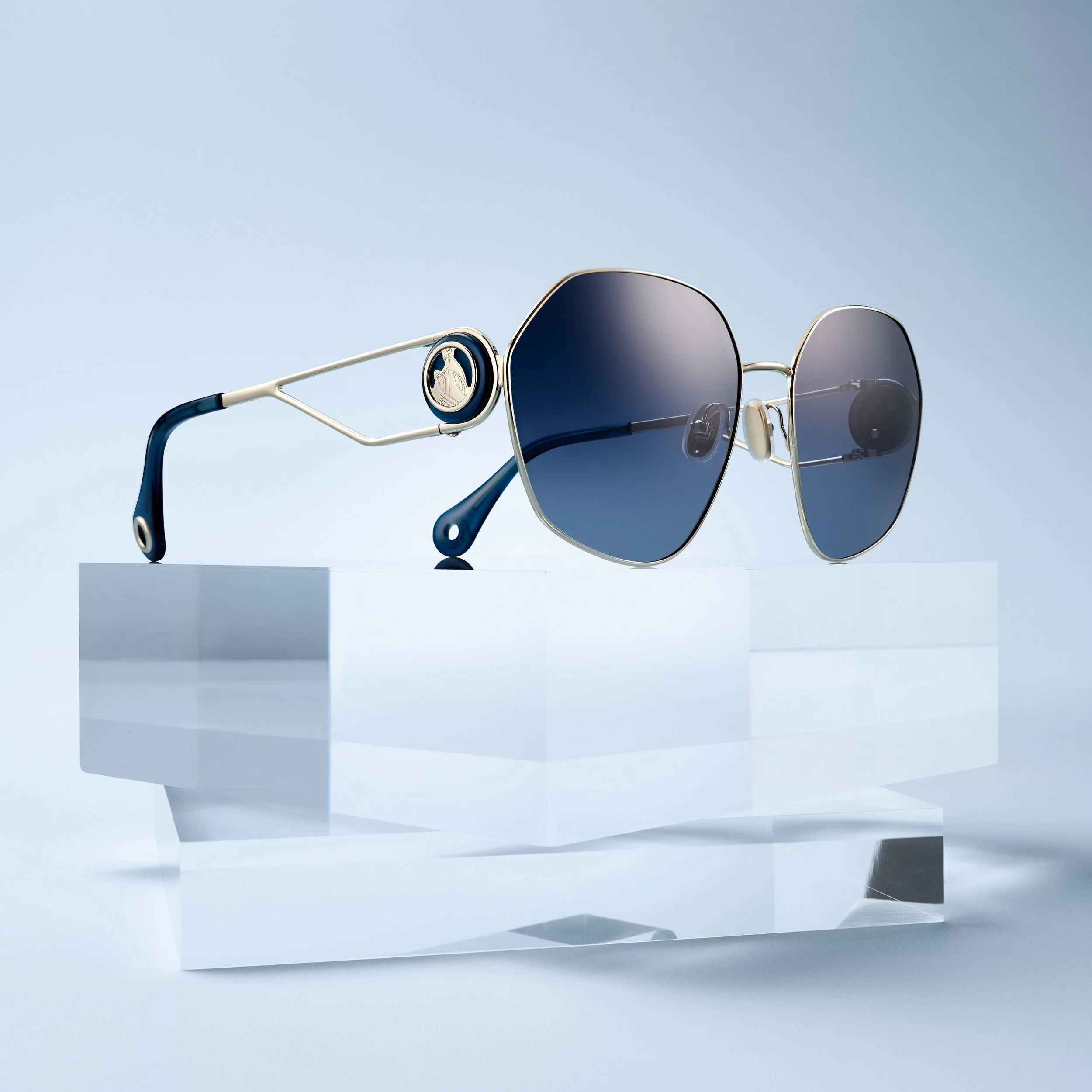 A pair of fashionable sunglasses with blue gradient lenses on a clear acrylic stand against a light blue background.