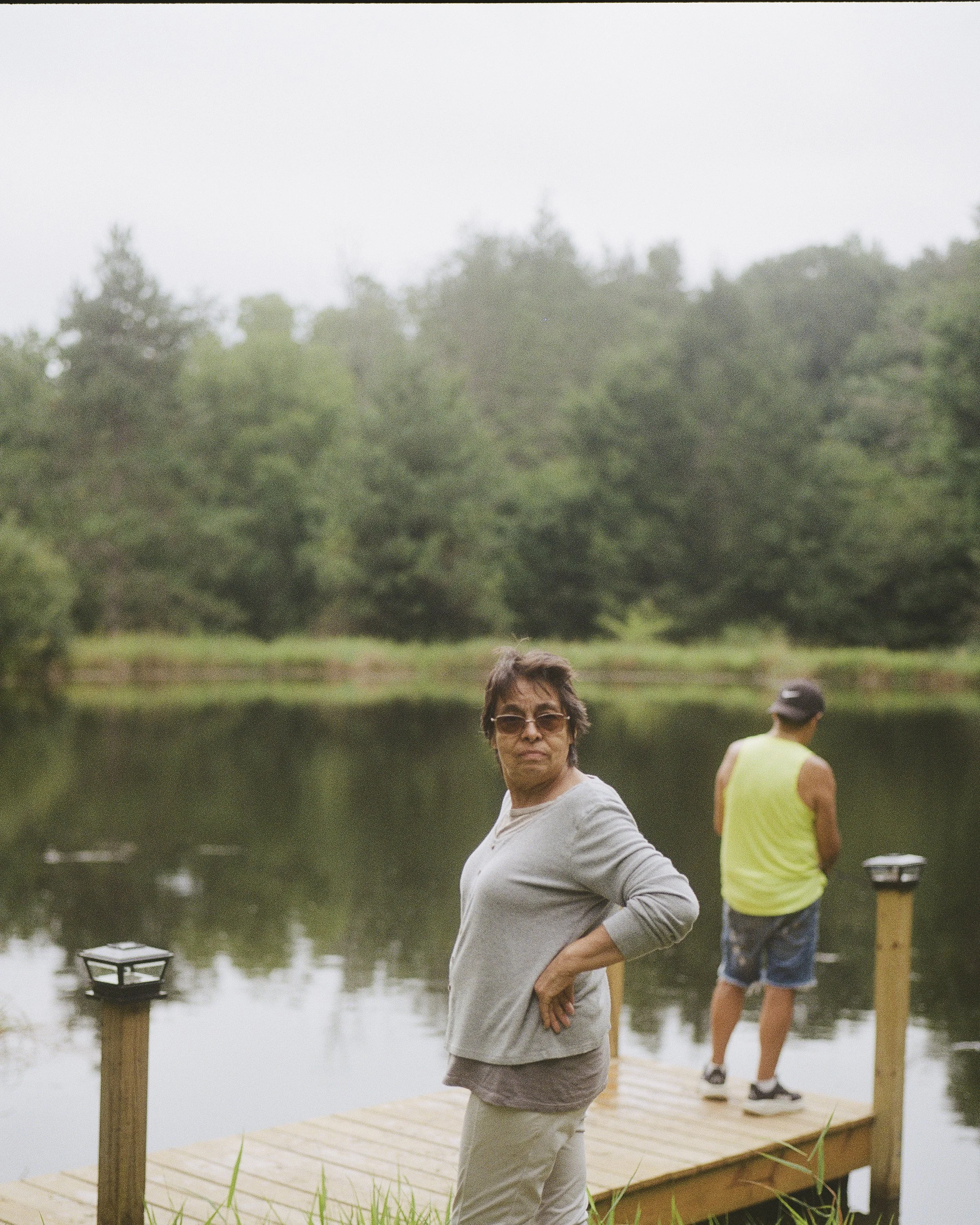 A woman standing by a lake on a wooden dock, with two men in the background near the water, one wearing a yellow vest and a cap, and the other in a shirt, with a treeline across the water and cloudy sky overhead.
