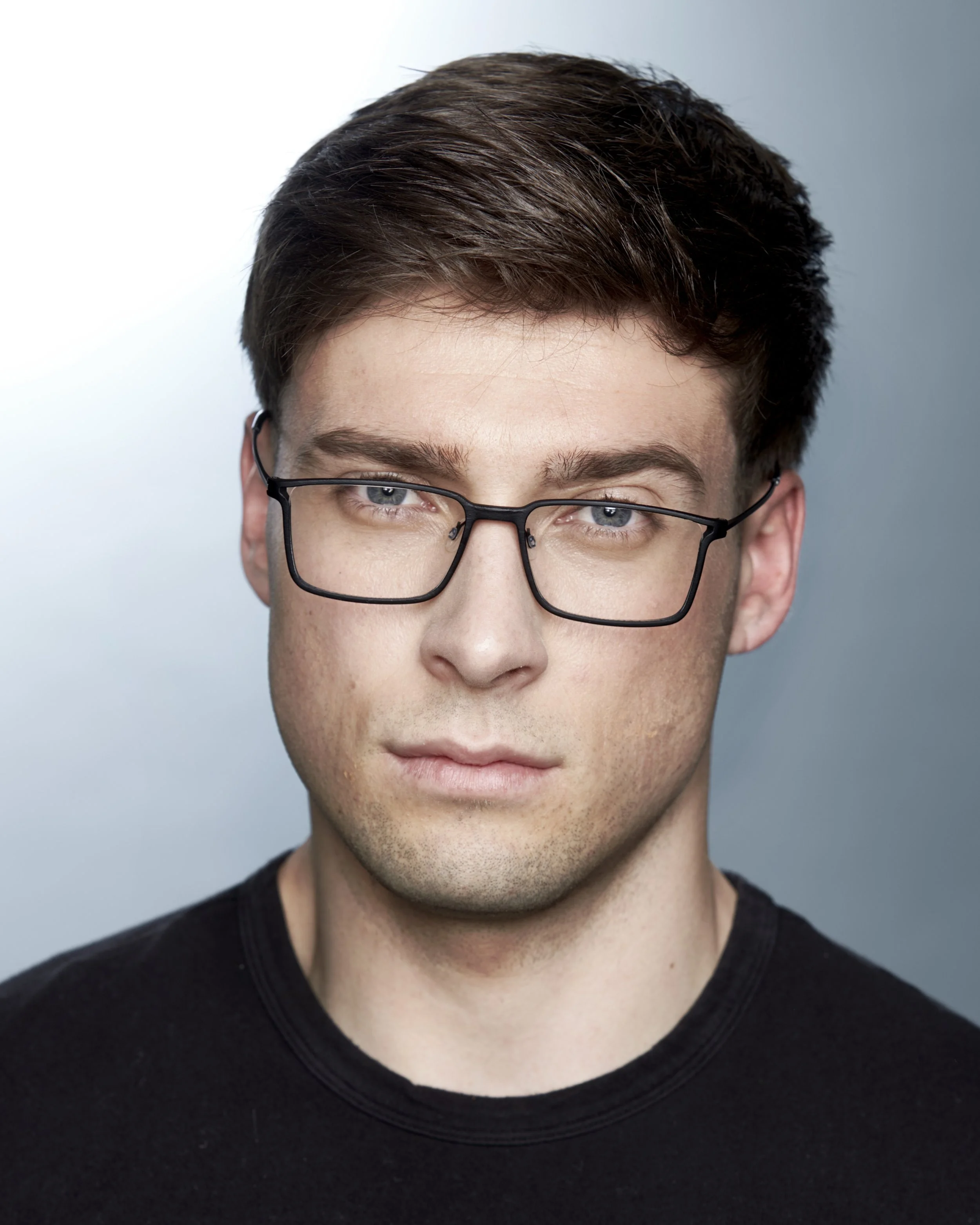 Close-up portrait of a young man with brown hair, blue eyes, wearing glasses, and a black shirt against a light background.