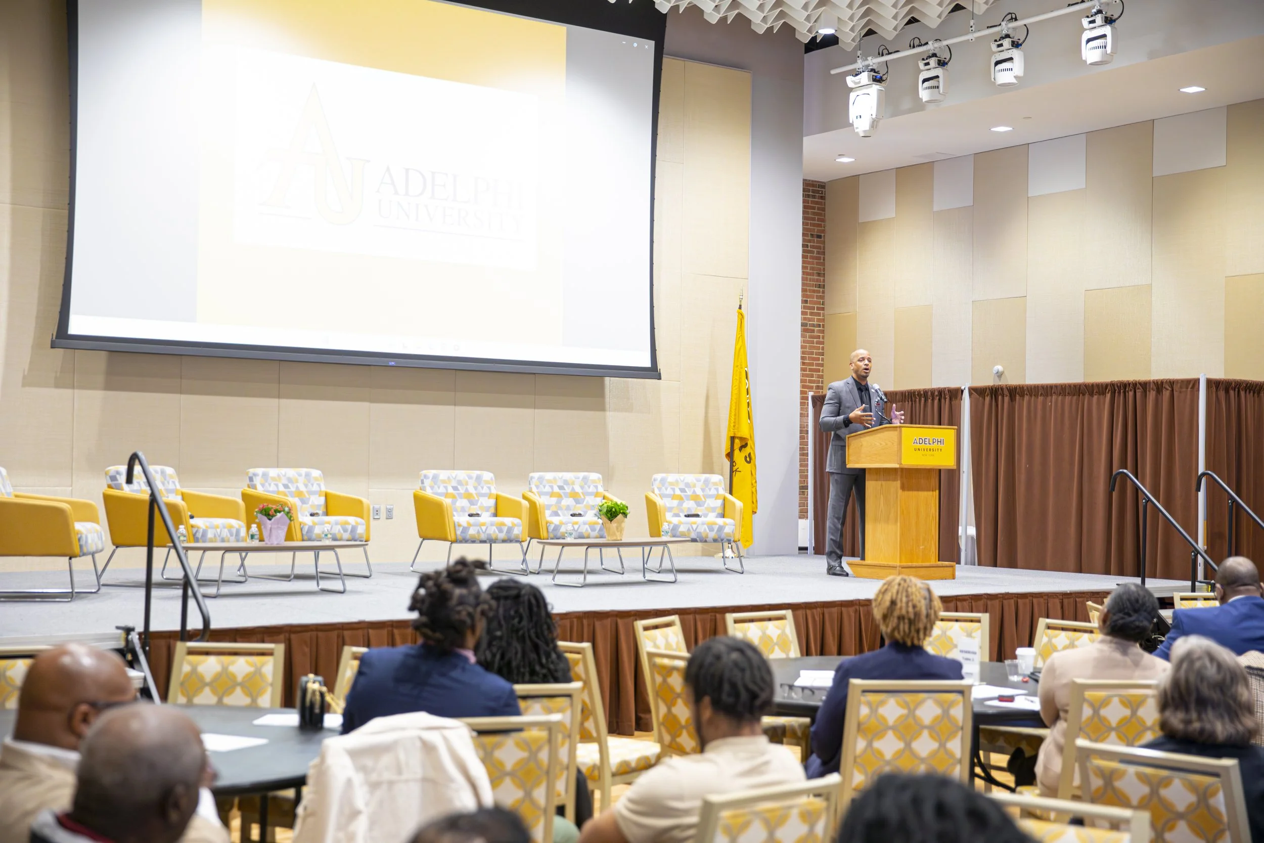 A speaker presenting at a conference stage with yellow chairs and a projection screen at Adelphi University, with attendees seated at round tables in the foreground.
