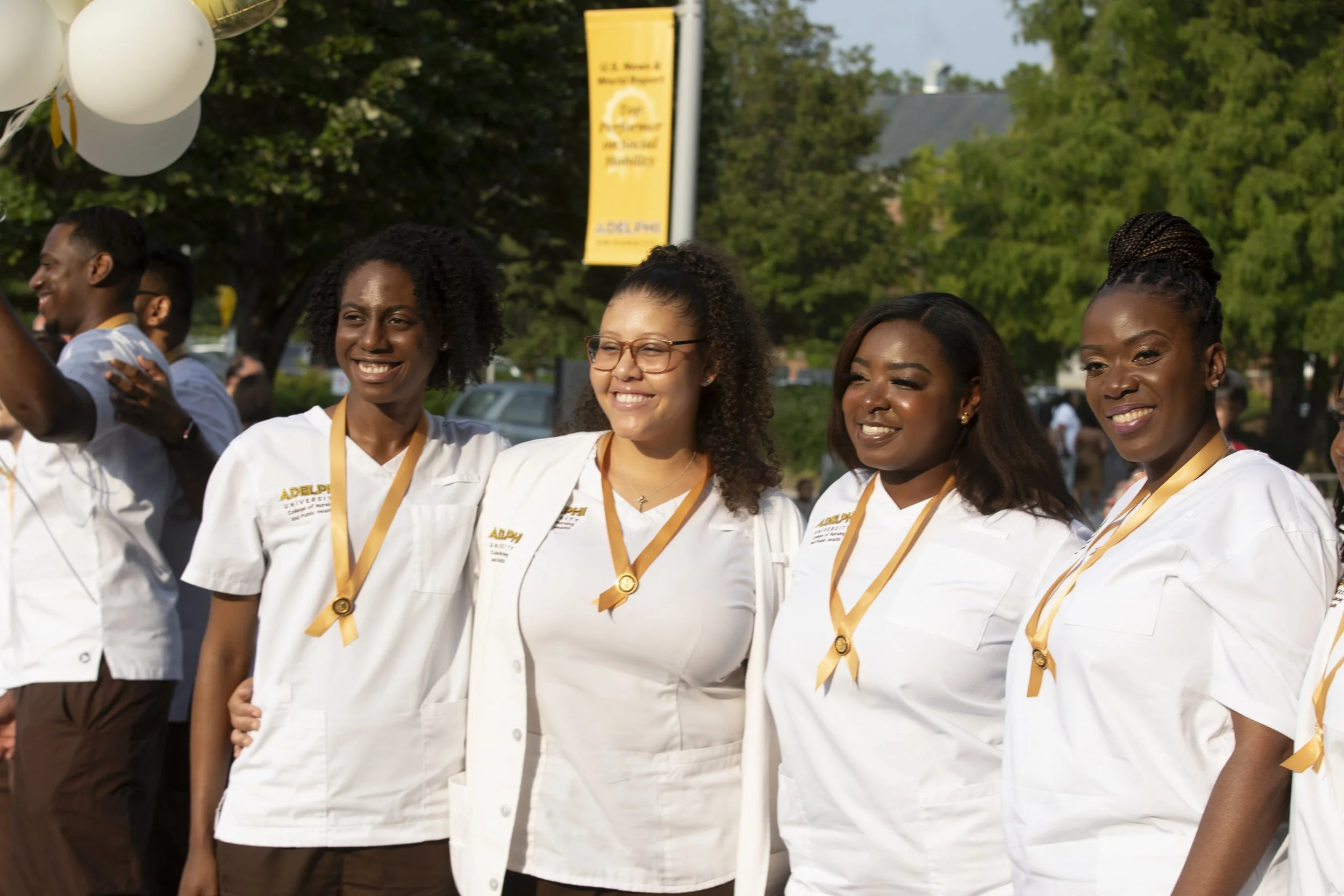 Group of women wearing white medical uniforms and gold medals, smiling outdoors during a celebration or graduation event.