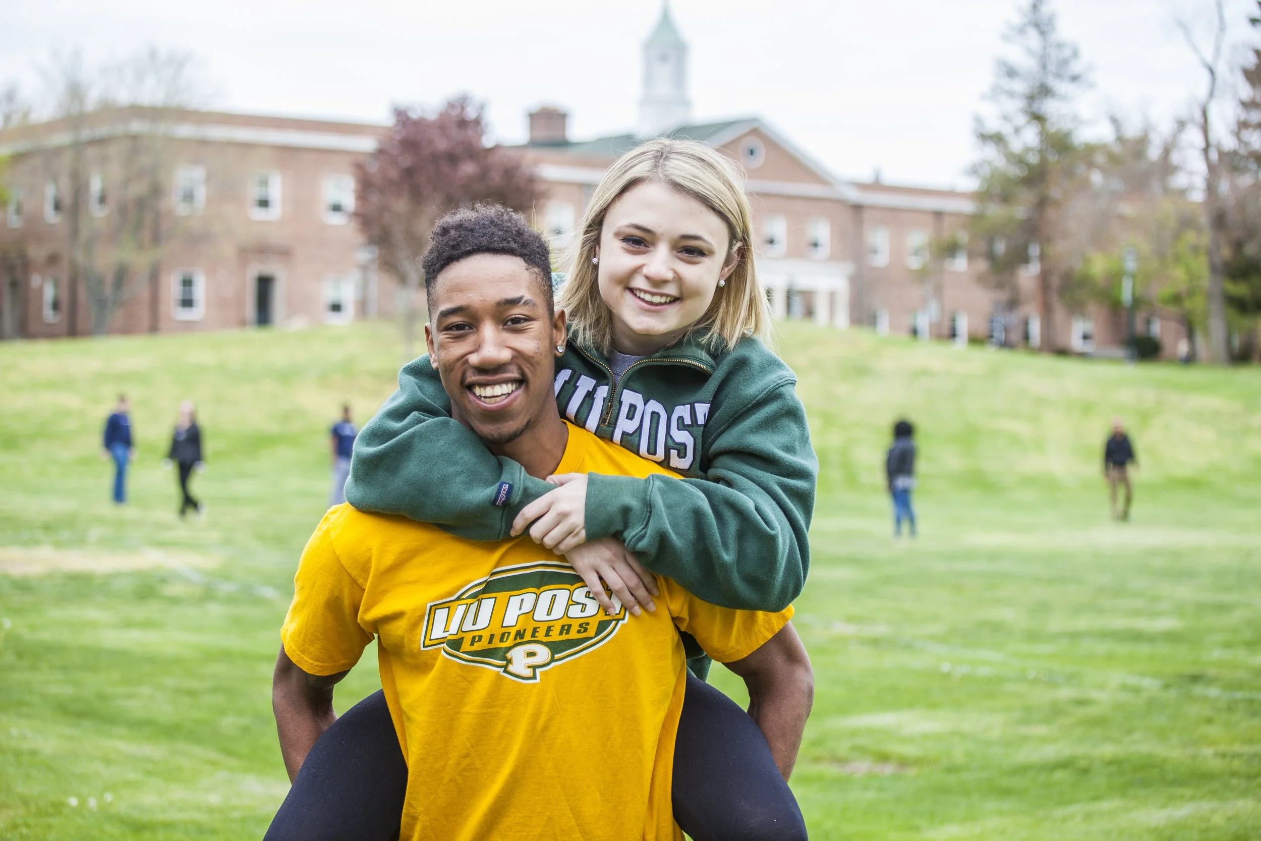 Two young people, a man and a woman, smiling outdoors on a grassy field, with a brick building and several people in the background.
