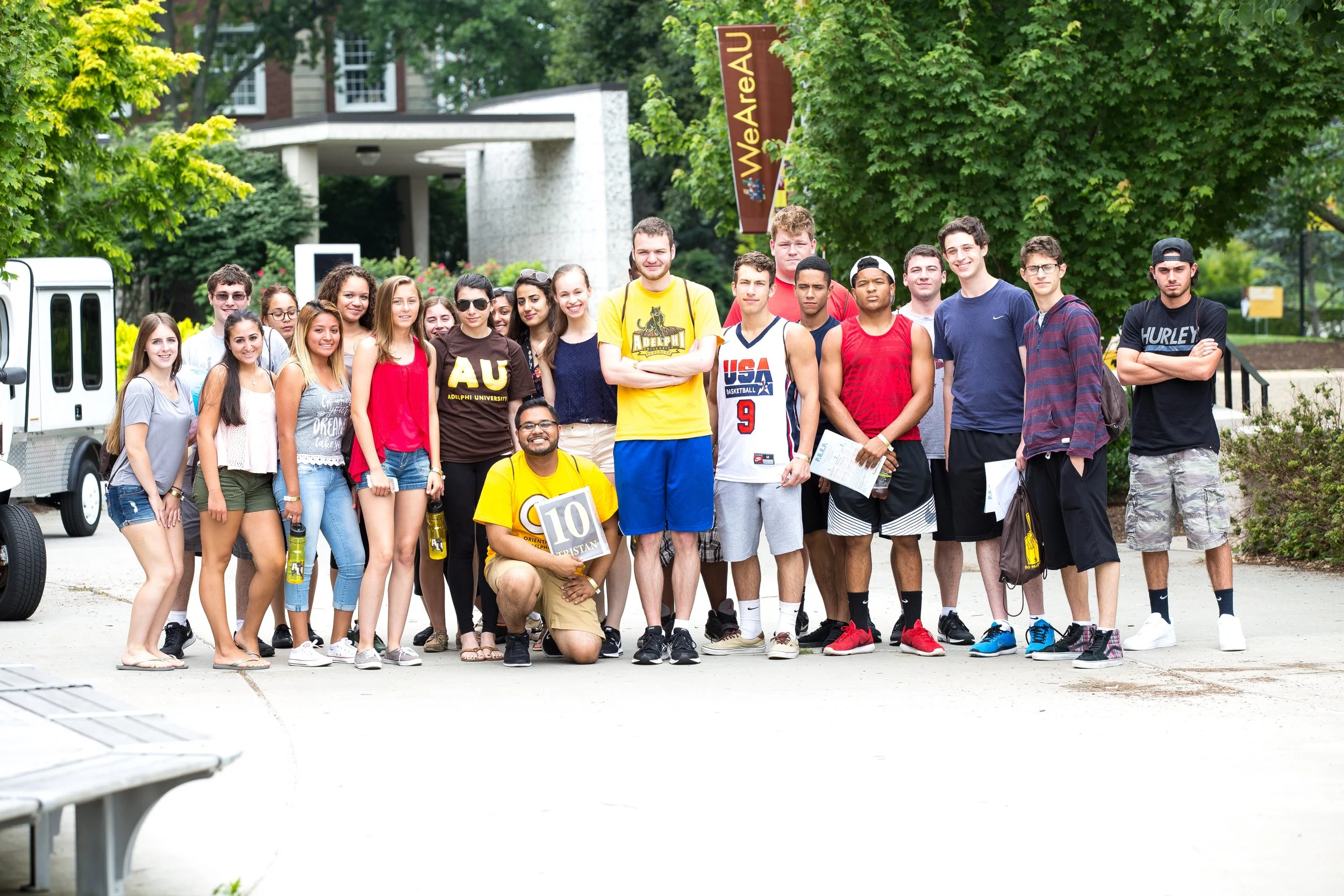 A group of young students standing outdoors on a university campus, with some holding papers and a student in a yellow shirt kneeling in front holding a small sign that says '10 Christian.' The background includes greenery, trees, and a campus sign t