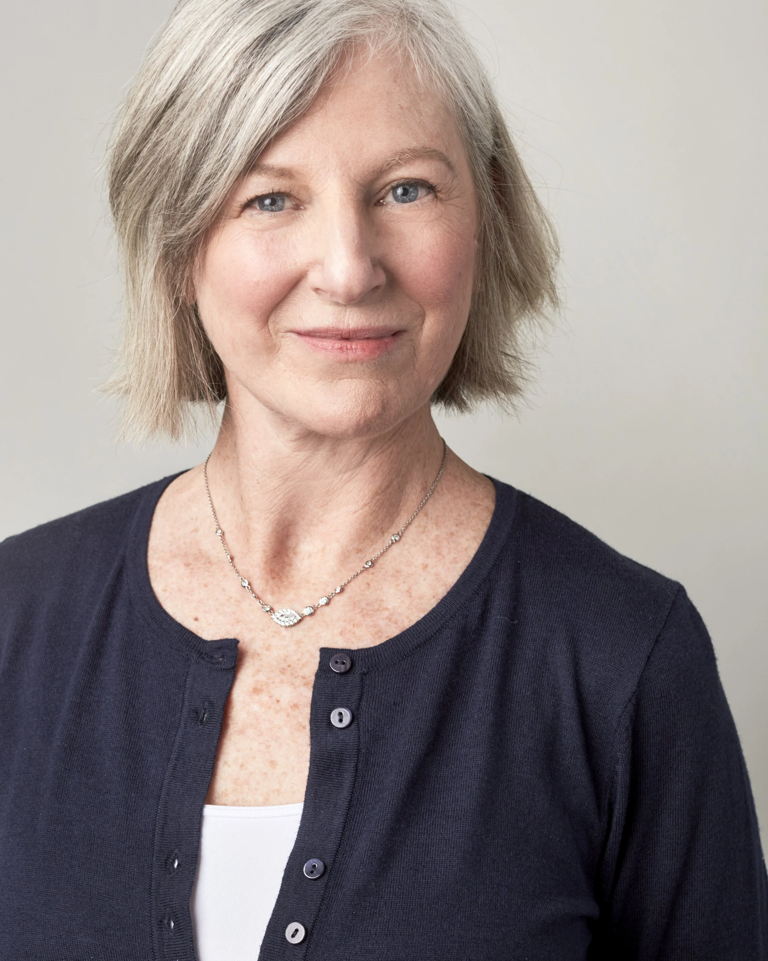 Portrait of a middle-aged woman with short gray hair, blue eyes, wearing a navy cardigan over a white top, and a silver necklace, against a plain light gray background.