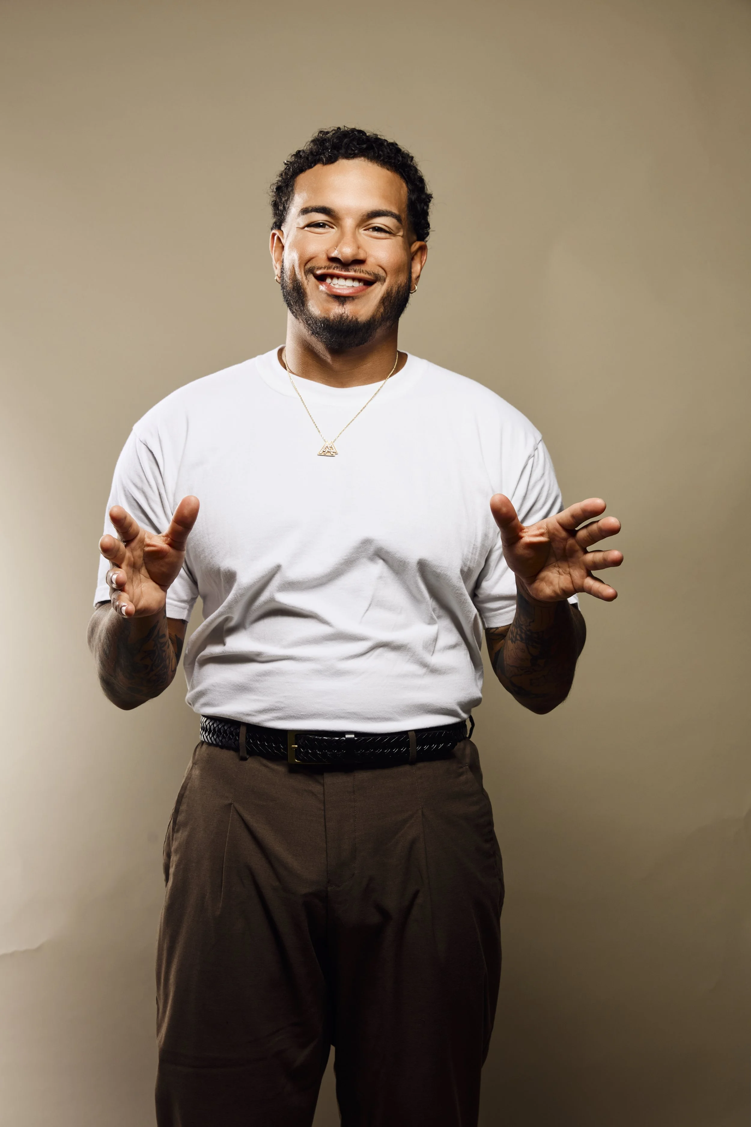 Smiling man with short curly hair, beard, and earrings, wearing a white t-shirt, a gold necklace, and brown pants, standing against a plain beige background.