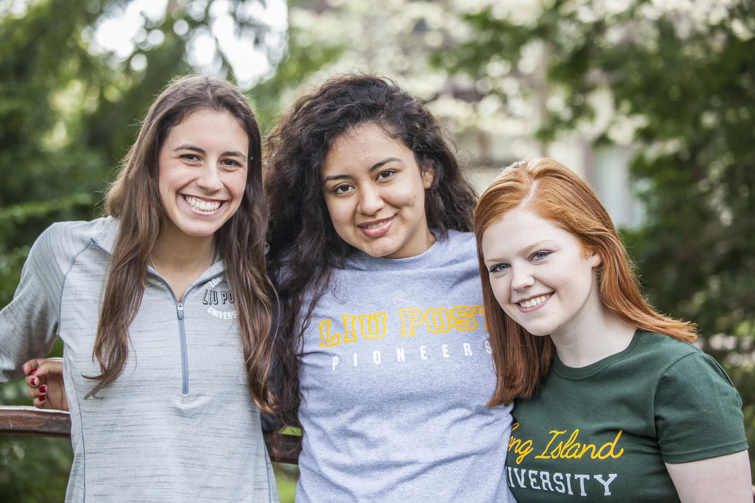 Three women smiling outdoors, one wearing a LIU Post sweatshirt, another wearing a LIU Post University shirt, and the third in a gray quarter-zip shirt, with a background of green trees.