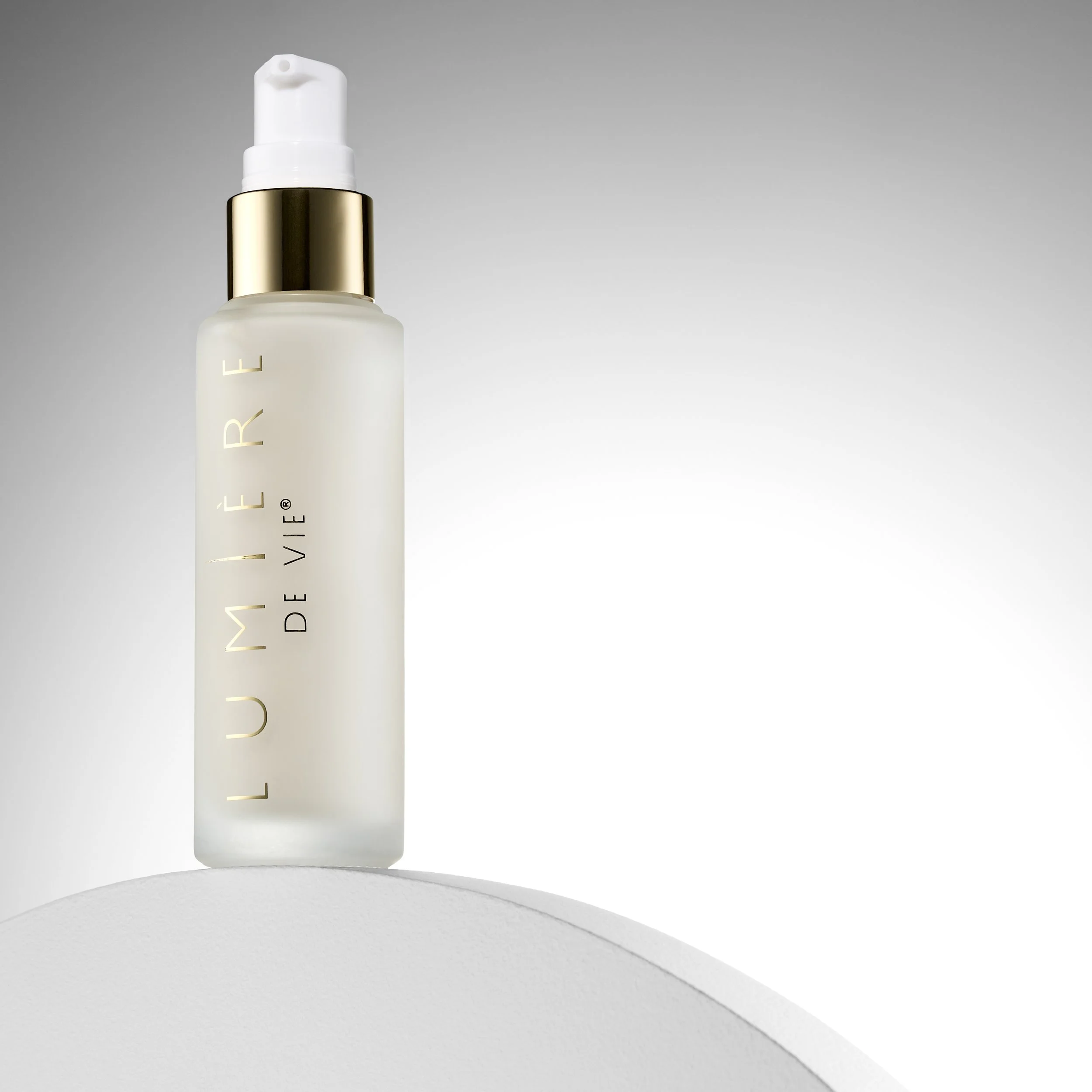 A frosted glass bottle of Lumière De Vie skincare product with a pump dispenser, gold and white accents, placed on a white curved surface against a gradient gray background.