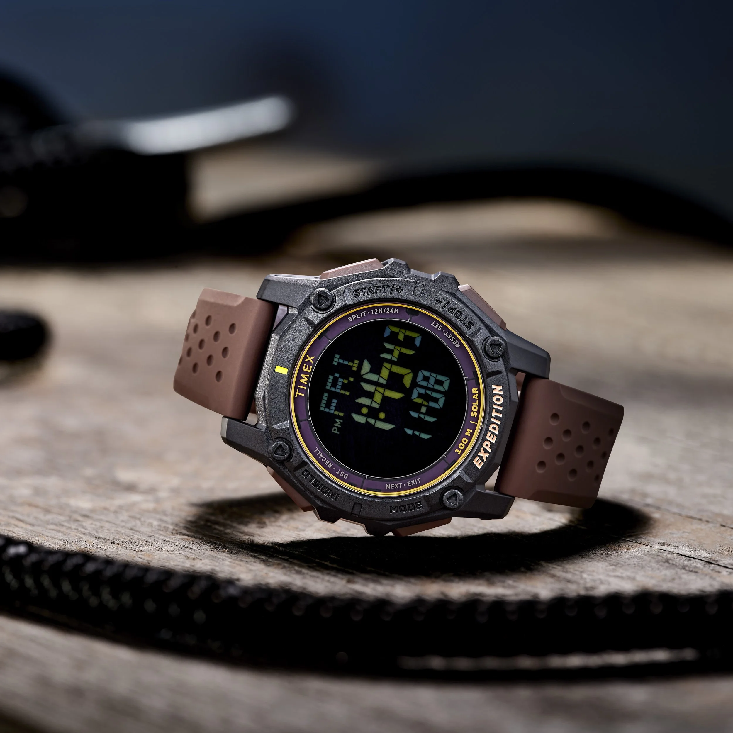 Digital wristwatch with brown strap and rugged black face displaying date, time, and additional features, placed on a wooden surface with blurred background.