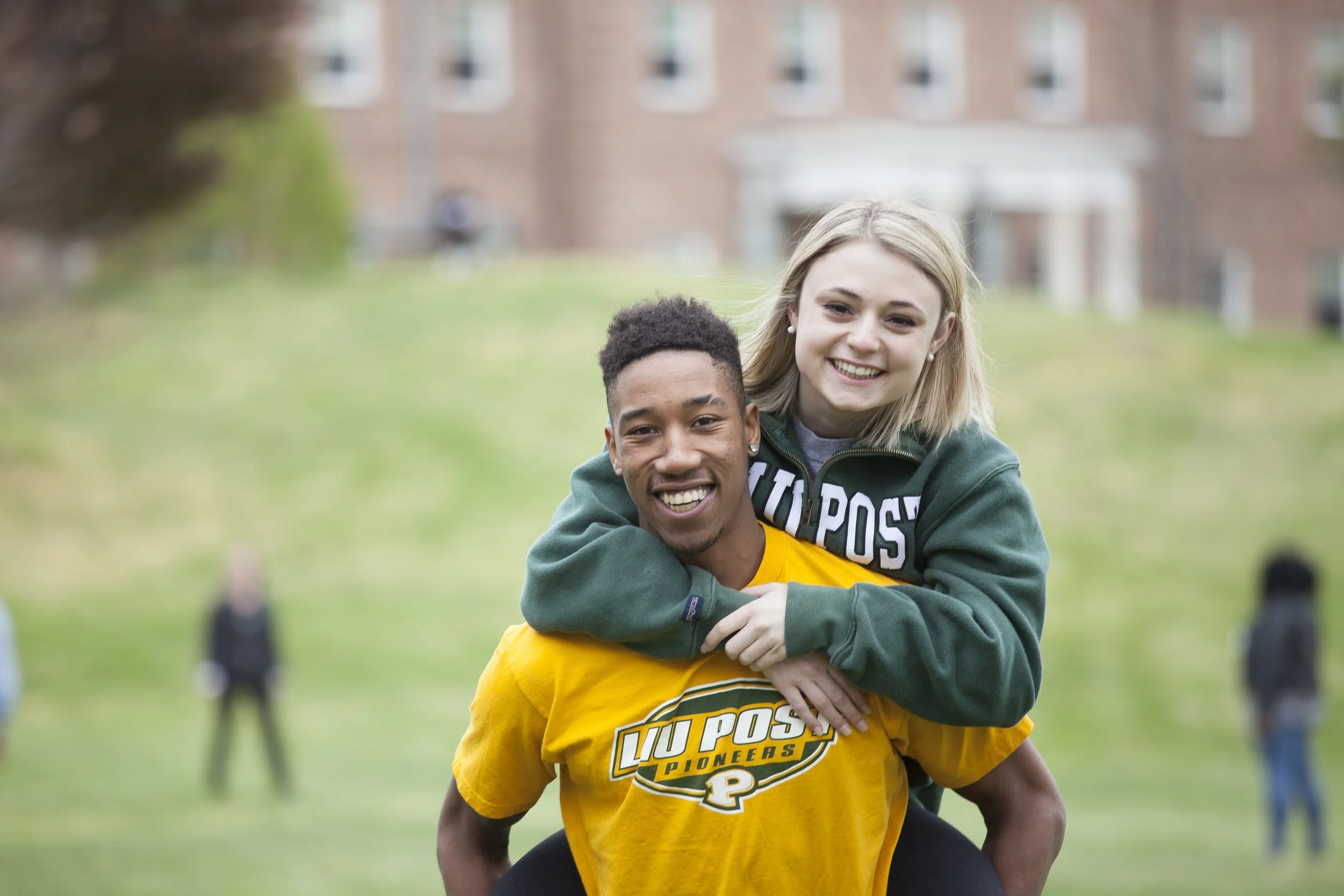 Smiling young man in a yellow T-shirt giving a piggyback ride to a smiling young woman in a green hoodie in a grassy outdoor setting with a building in the background.
