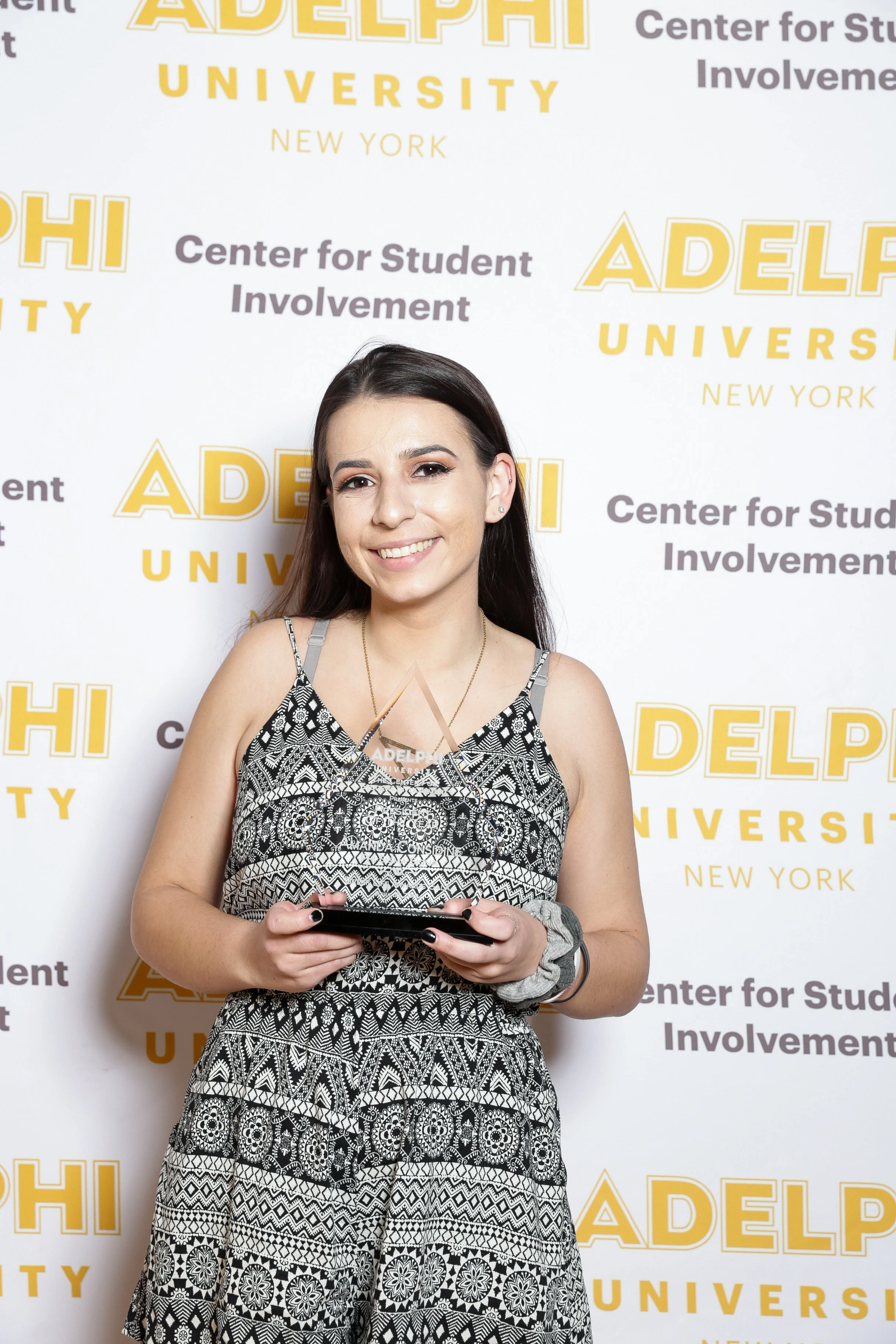 A young woman with long dark hair and a smile, standing in front of a backdrop with the text "ADELPI UNIVERSITY NEW YORK" and "Center for Student Involvement." She is wearing a black and white patterned dress, holding a phone, and has a gray scrunchi