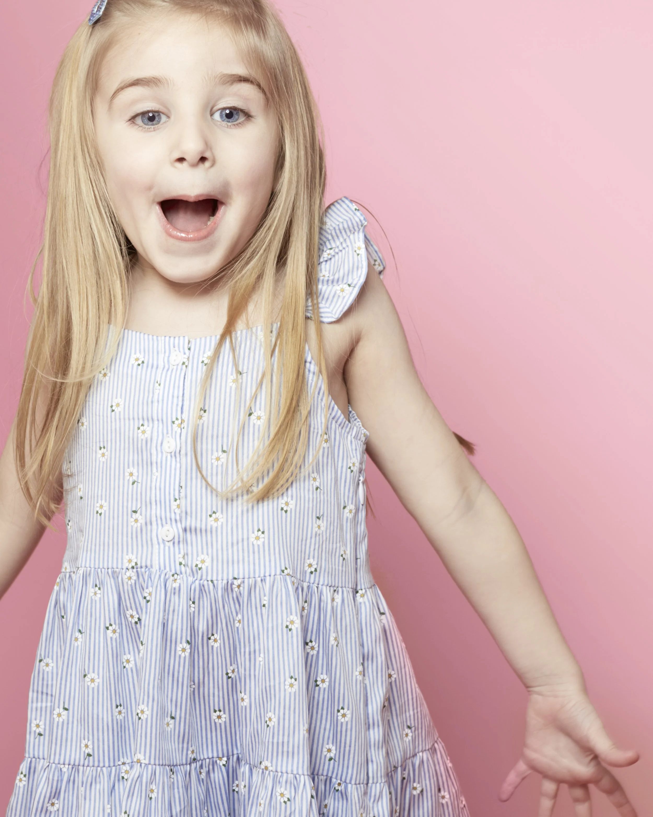 A young girl with long blonde hair, wearing a blue and white striped dress with small flower patterns, posing with her mouth open and an expression of surprise or excitement, against a pink background.