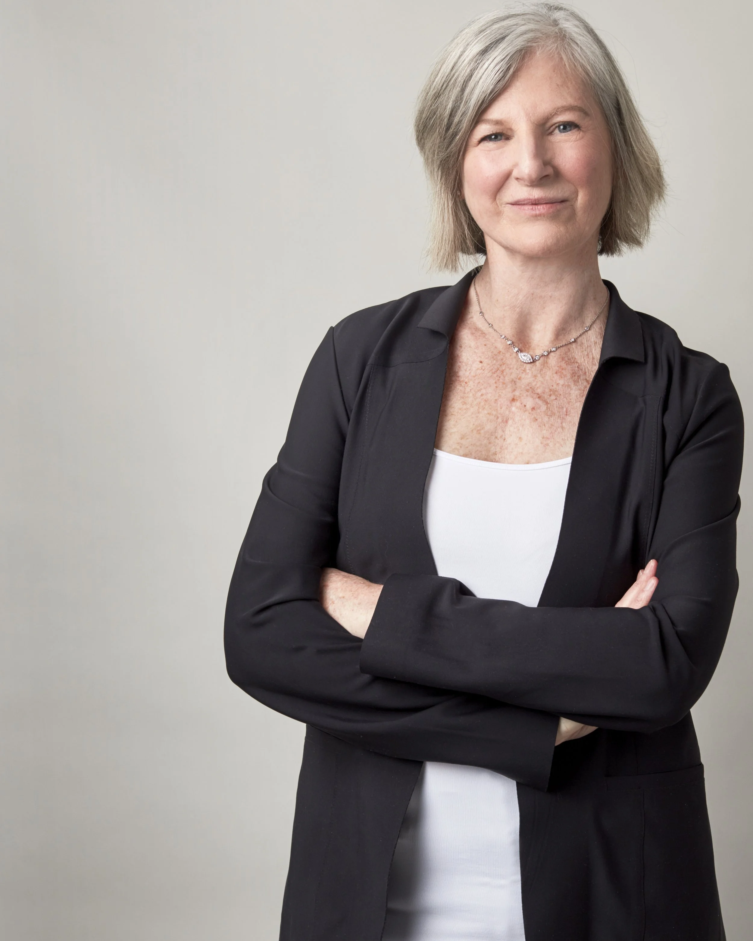 Portrait of a middle-aged woman with short gray hair, wearing a black blazer over a white top, crossing her arms, standing against a plain gray background.