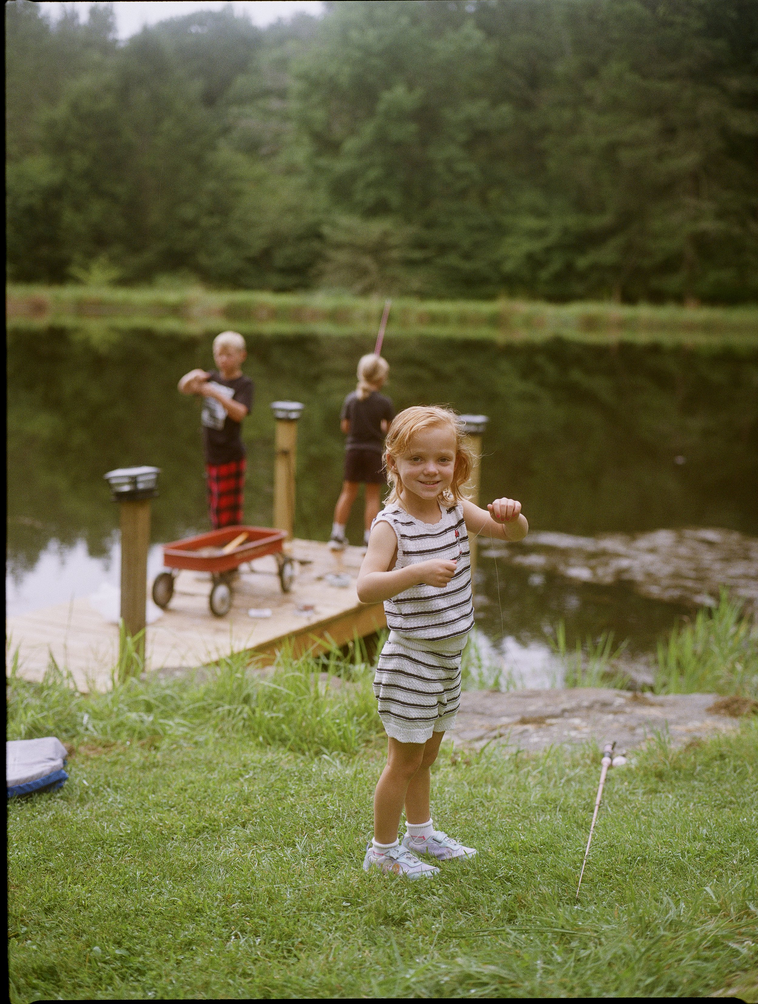 A young girl standing on a grassy area near a small pond, holding a fishing rod, smiling, with two boys in the background near the water, surrounded by trees.