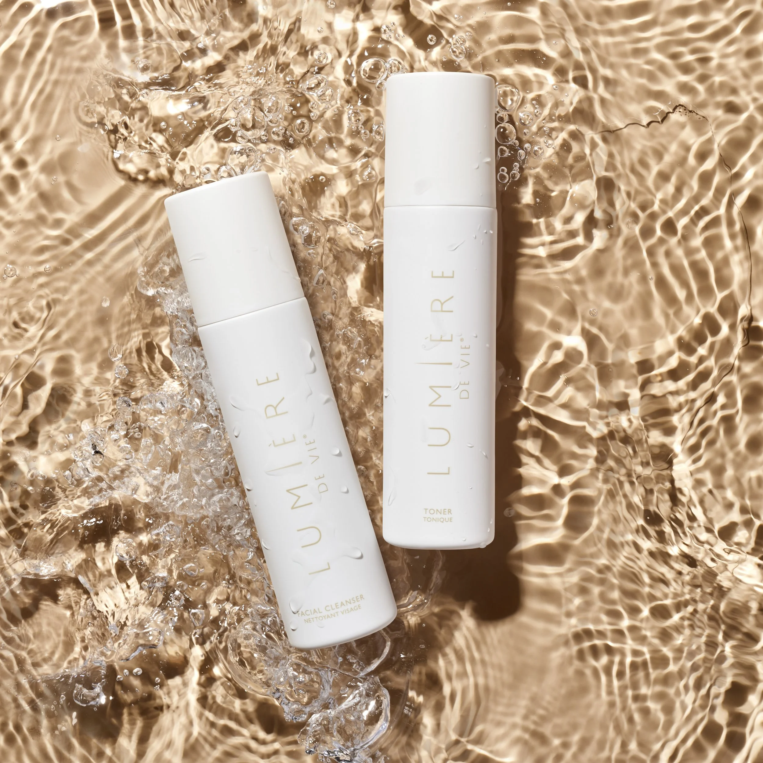 Two white skincare bottles, labeled 'LUMIÈRE de VIE', are lying on warm water with ripples and bubbles, one labeled 'Facial Cleanser' and the other 'Toner'.