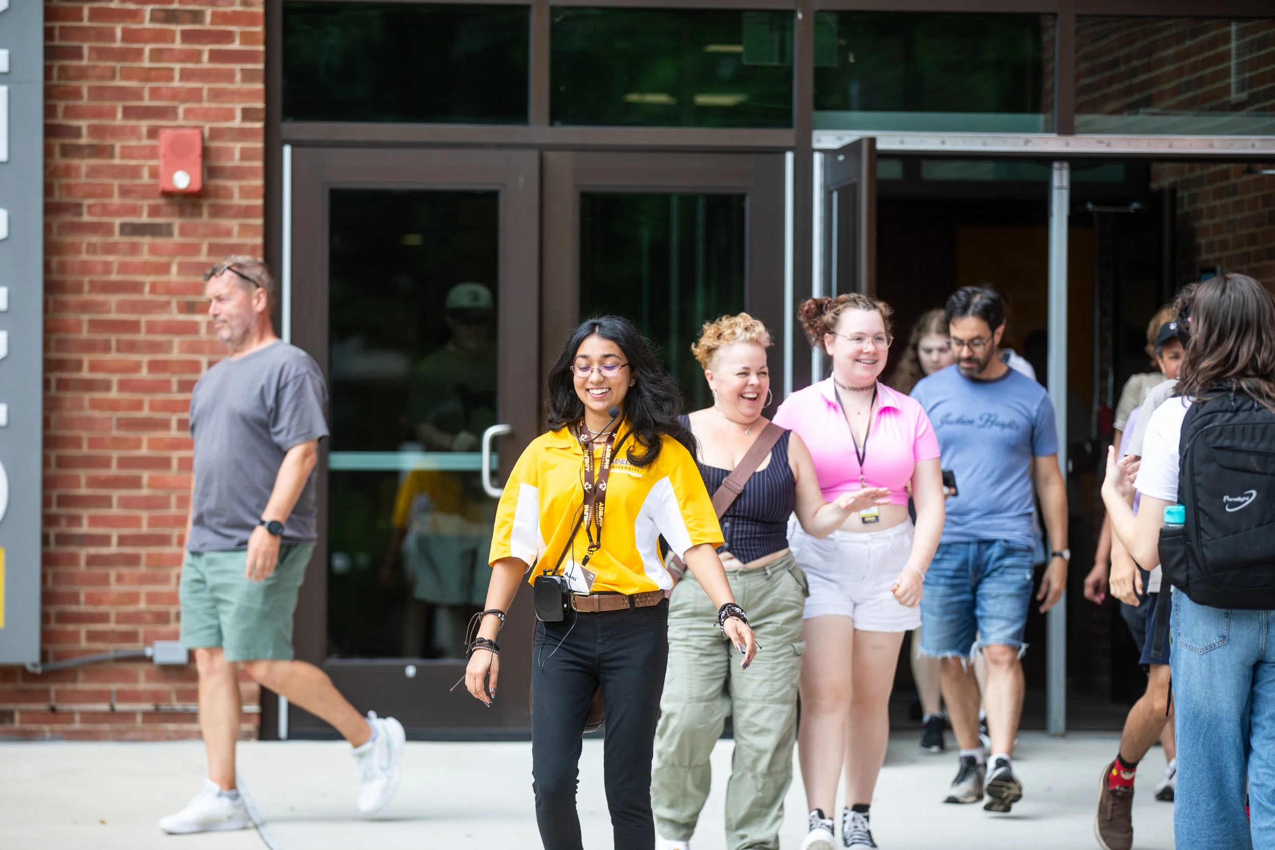 Group of diverse young adults walking out of a building, smiling, with a woman in a yellow shirt leading the group.