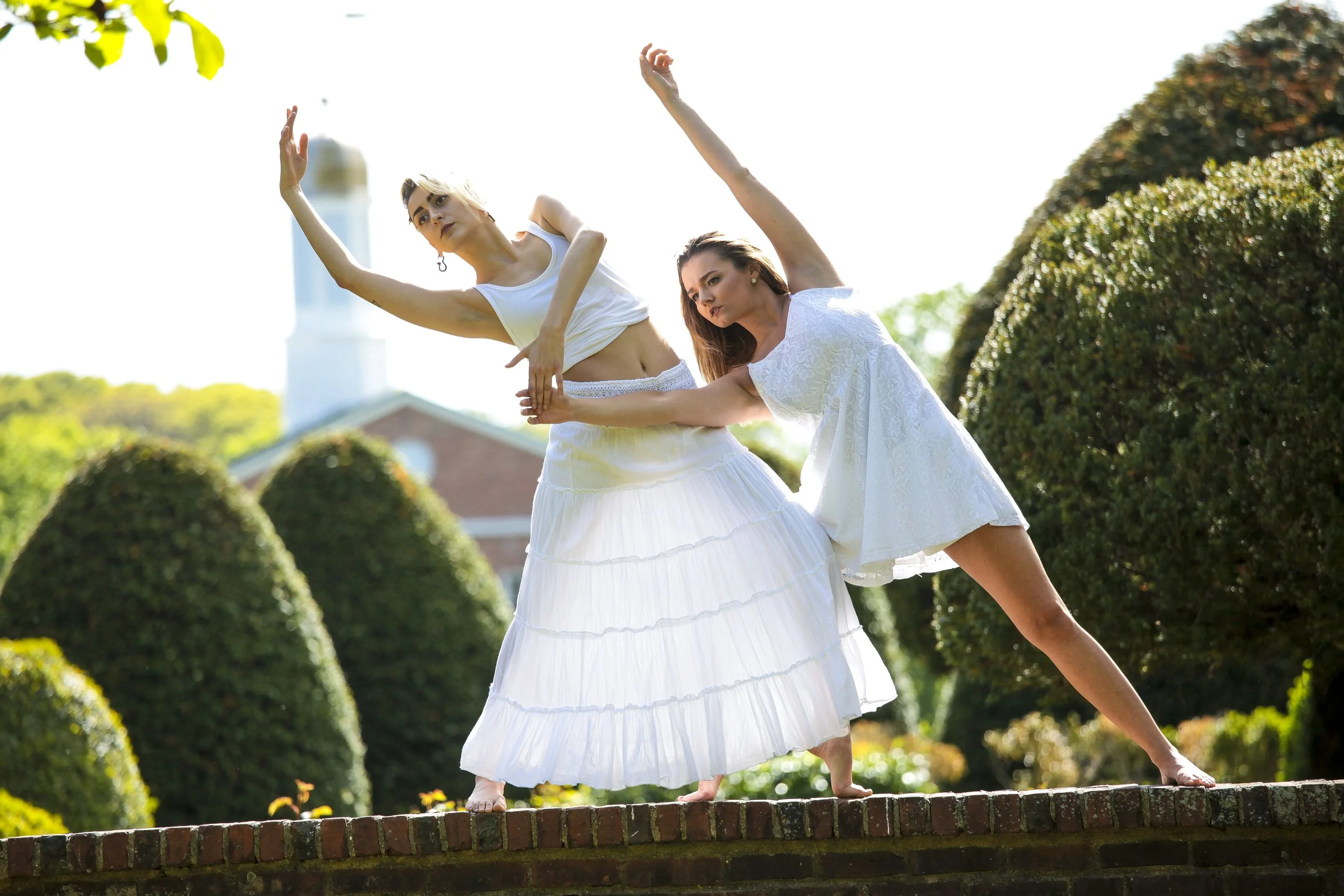 Two women in white dresses performing dance poses outdoors on a brick ledge, with green bushes and a church steeple in the background.