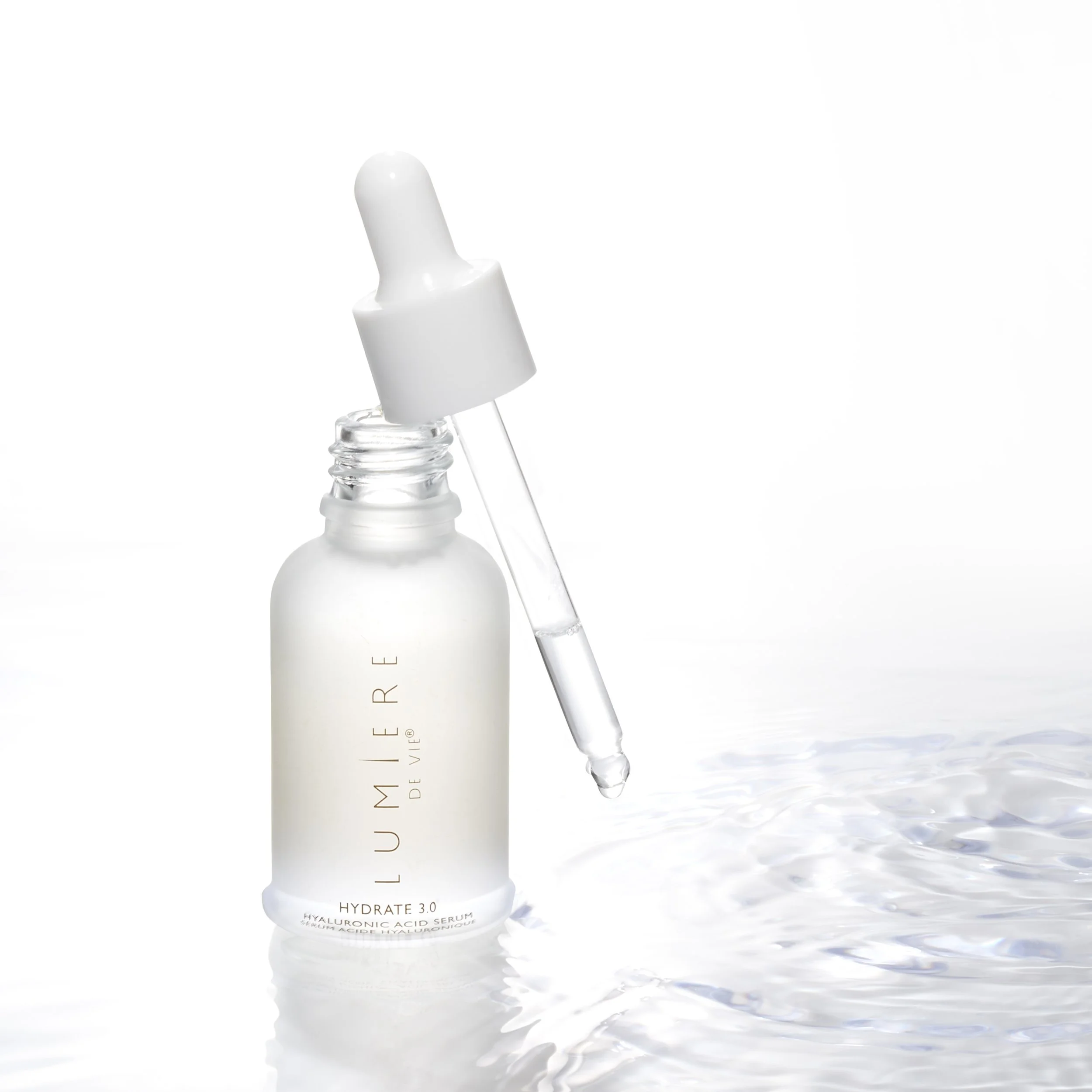 A white dropper bottle labeled 'LUMIERE' with a glass pipette and a drop of serum, placed on a reflective surface with water ripples.