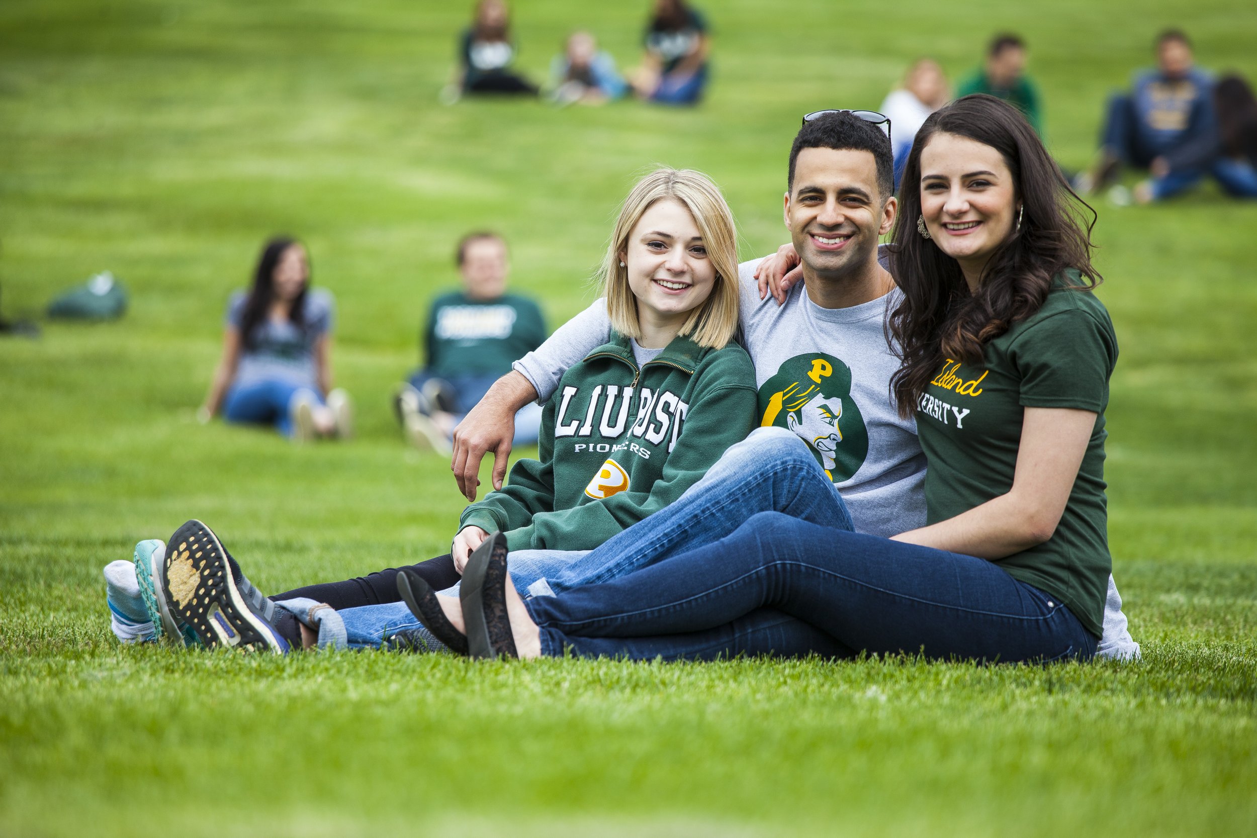 Three smiling young adults sitting on grass outdoors, with a group of people blurred in the background. They are wearing casual clothing, including hoodies and t-shirts with university logos.