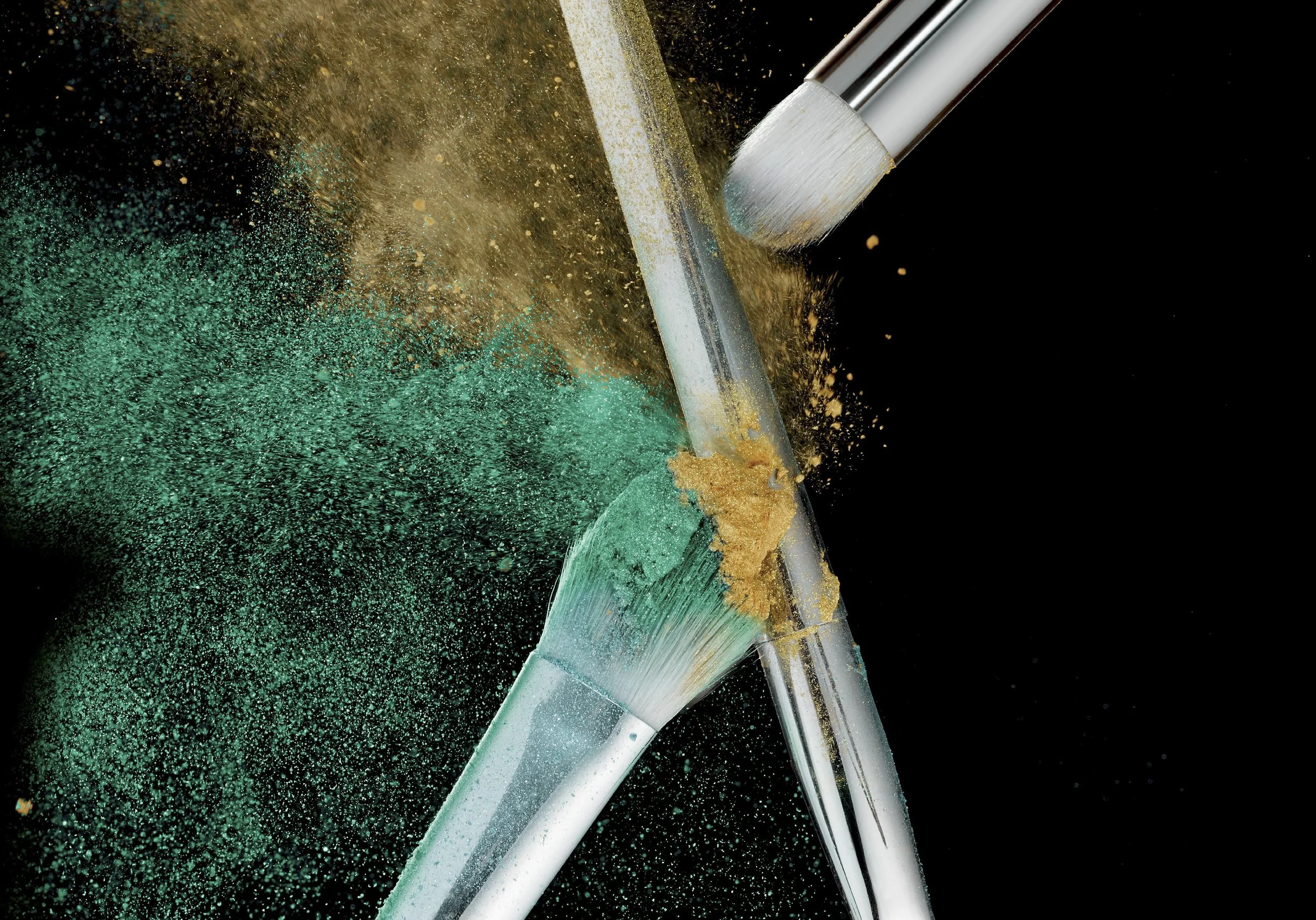 Makeup brushes breaking, with green and gold powder dispersing into the air against a black background.