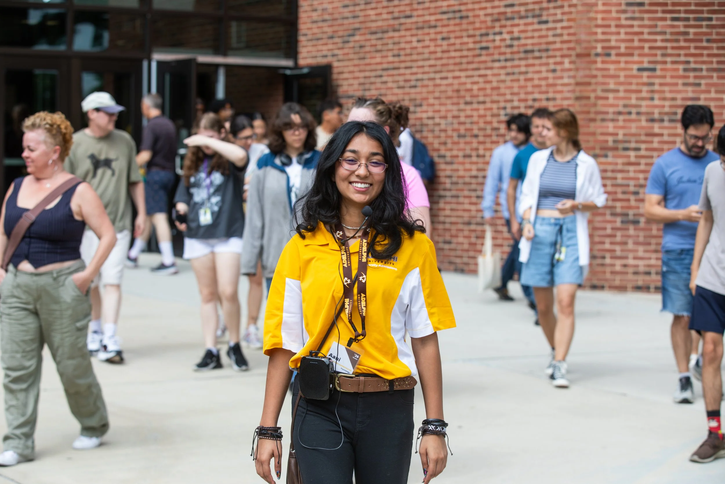 A young woman with glasses, long dark hair, and a yellow and white shirt smiling while walking outside among a crowd of people near a brick building.
