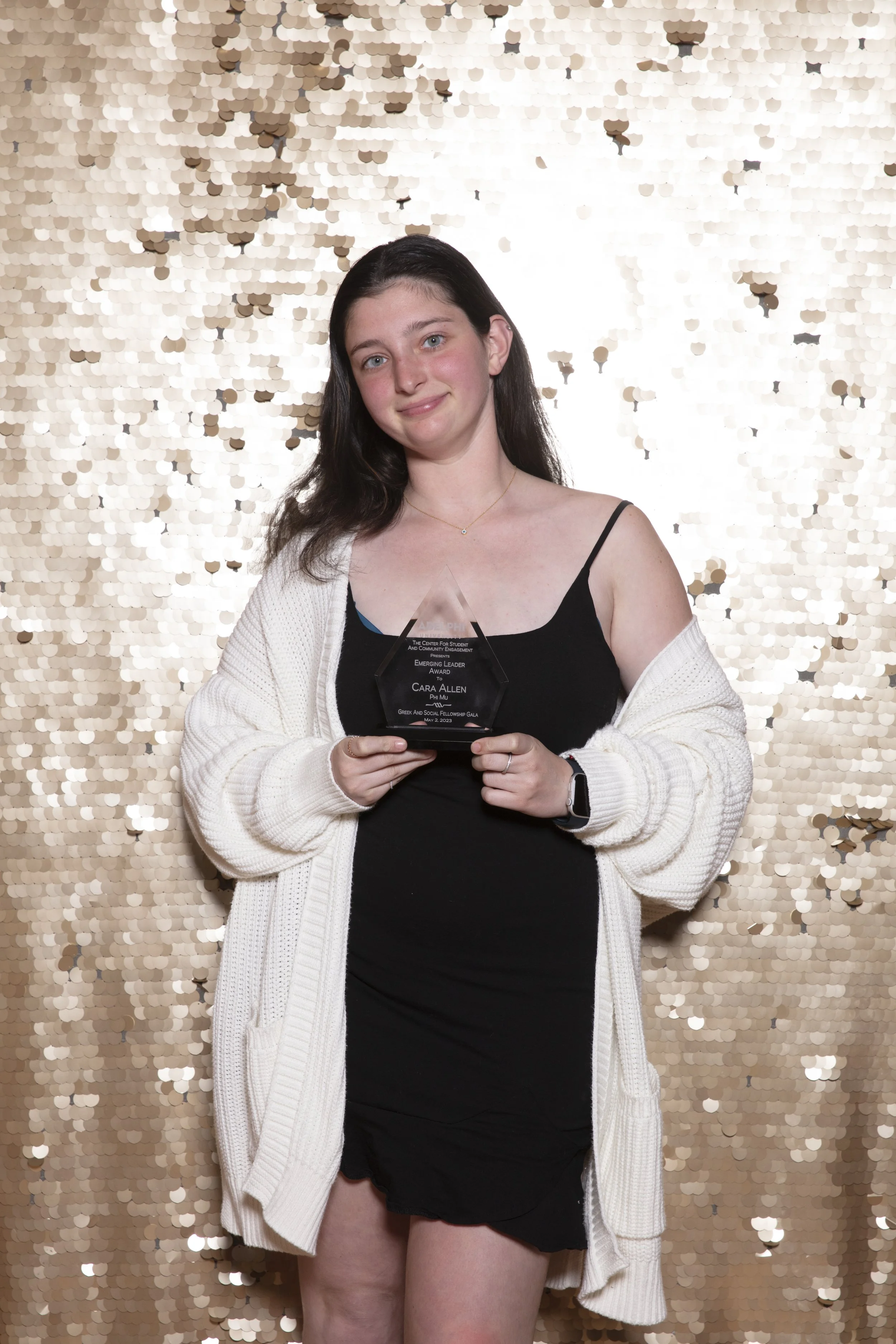 Young woman with long dark hair wearing a black dress and white cardigan, holding an award, standing in front of a gold sequin backdrop.