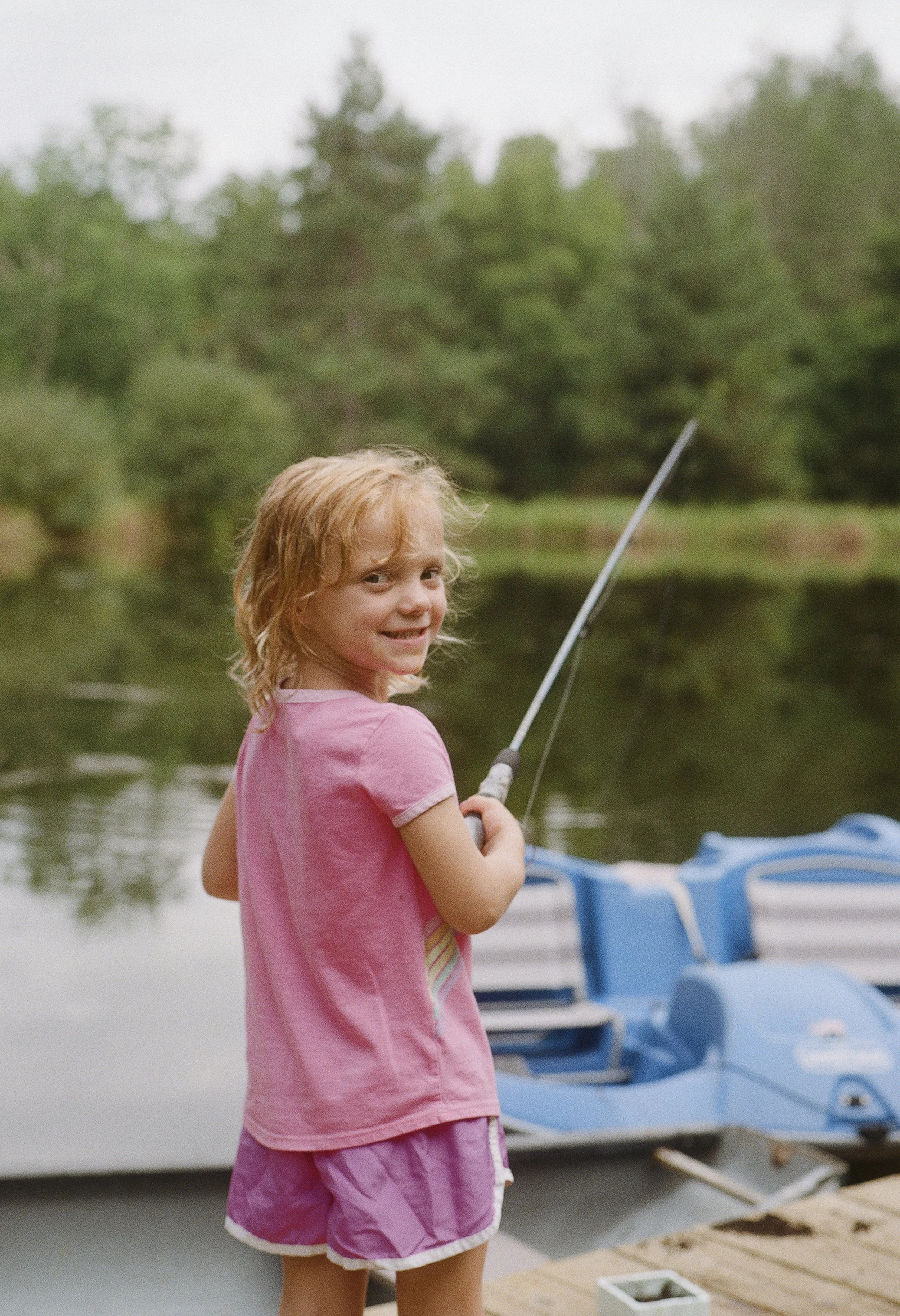 A young girl with blonde hair wearing a pink shirt and pink shorts, standing near a boat on a dock, holding a fishing rod, with trees and water in the background.