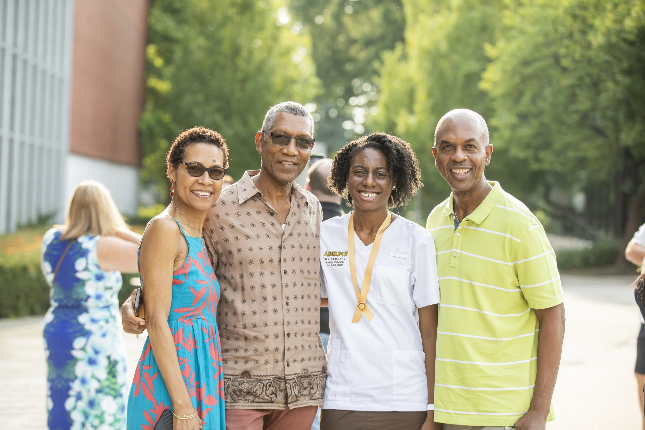 Four smiling adults standing outdoors in sunny weather, with trees and other people in the background.