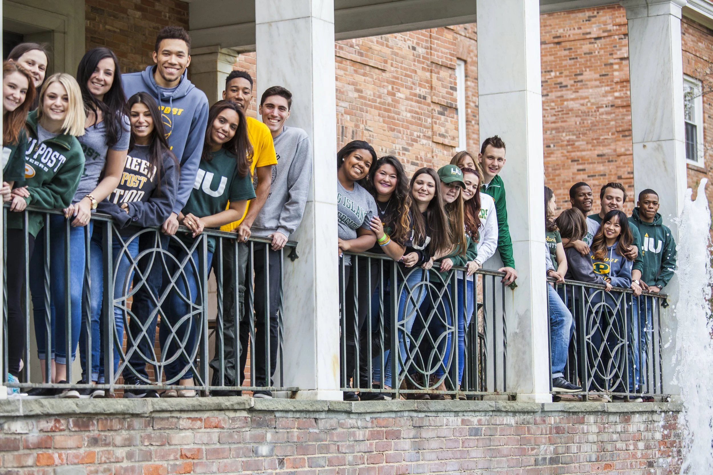 Group of diverse college students leaning on a decorative railing on a porch with brick walls, smiling for the photo.