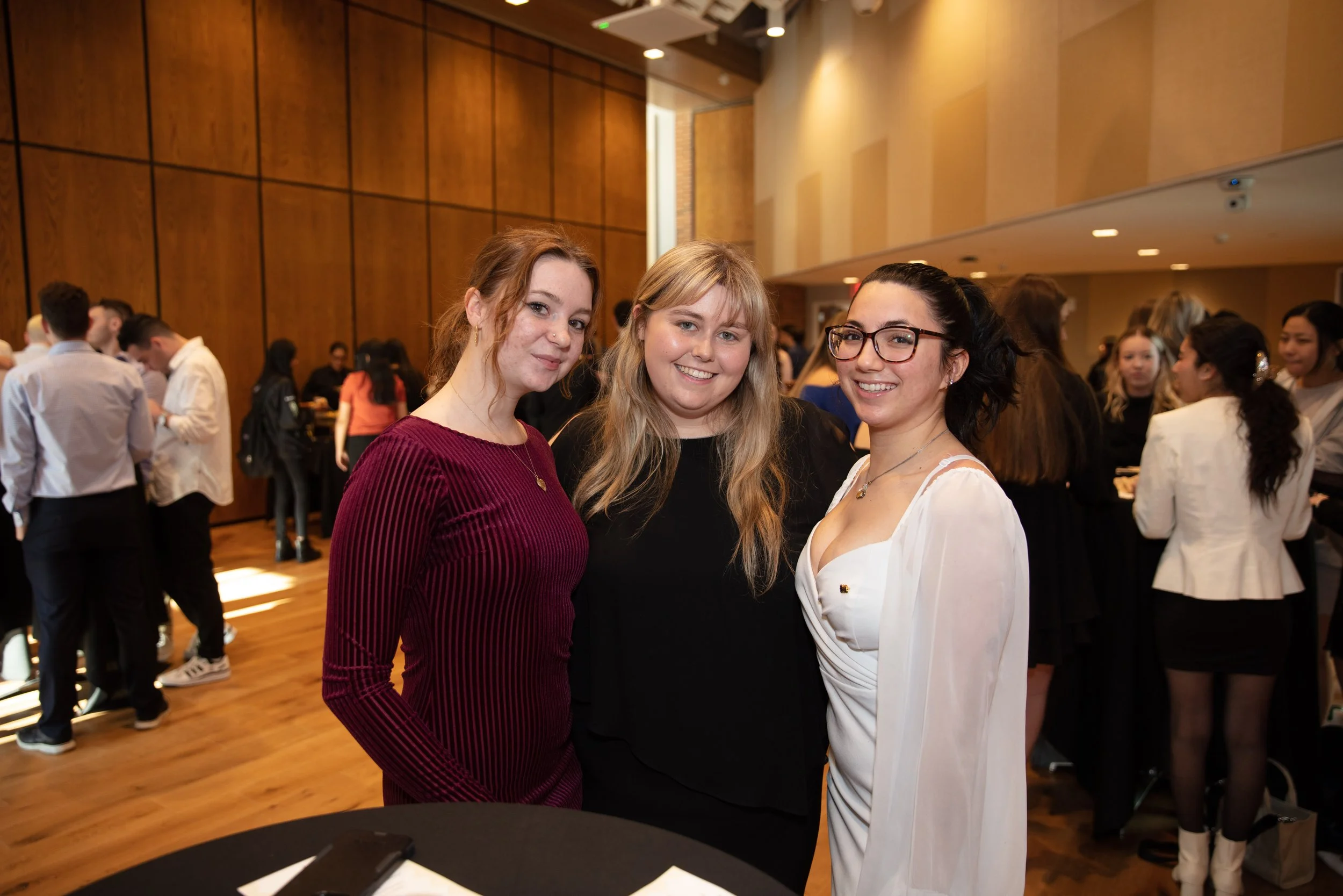Three women smiling at an indoor event with a crowd in the background.