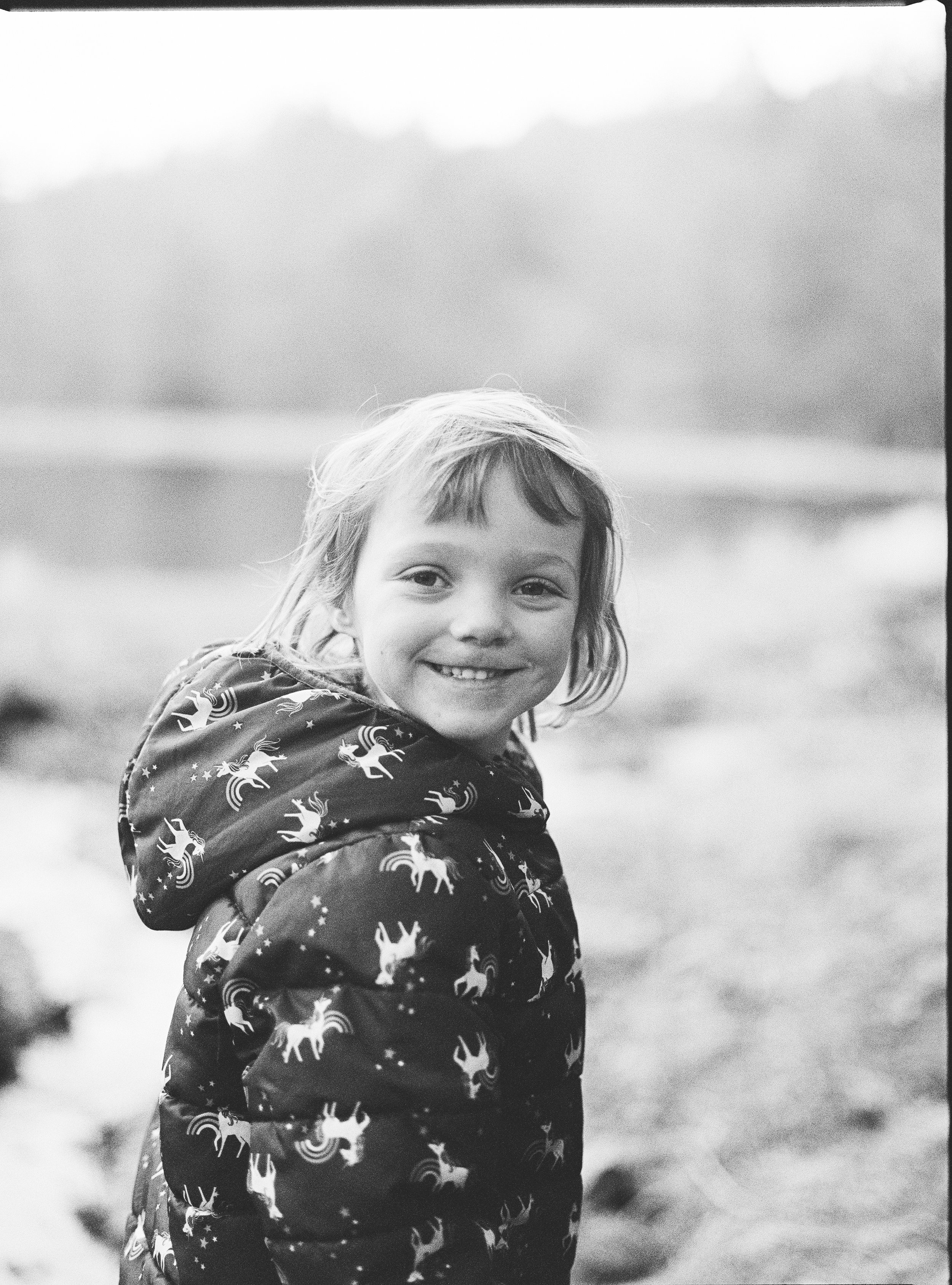 Black and white photo of a young girl with a bright smile, wearing a jacket with unicorn and star patterns, outdoors with a blurred natural background and cloudy sky.