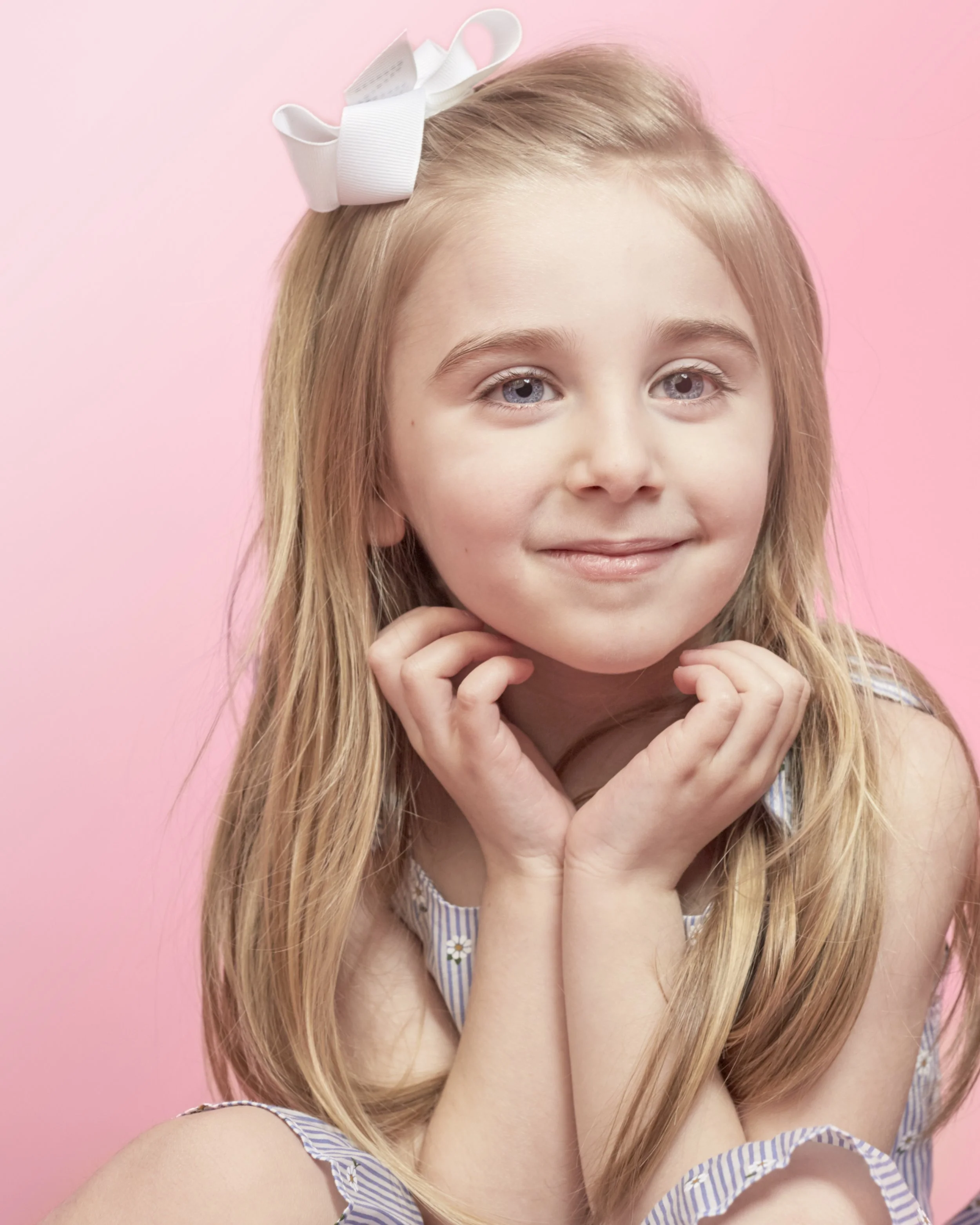 A young girl with long blonde hair, blue eyes, and a white bow in her hair, smiling softly against a pink background.
