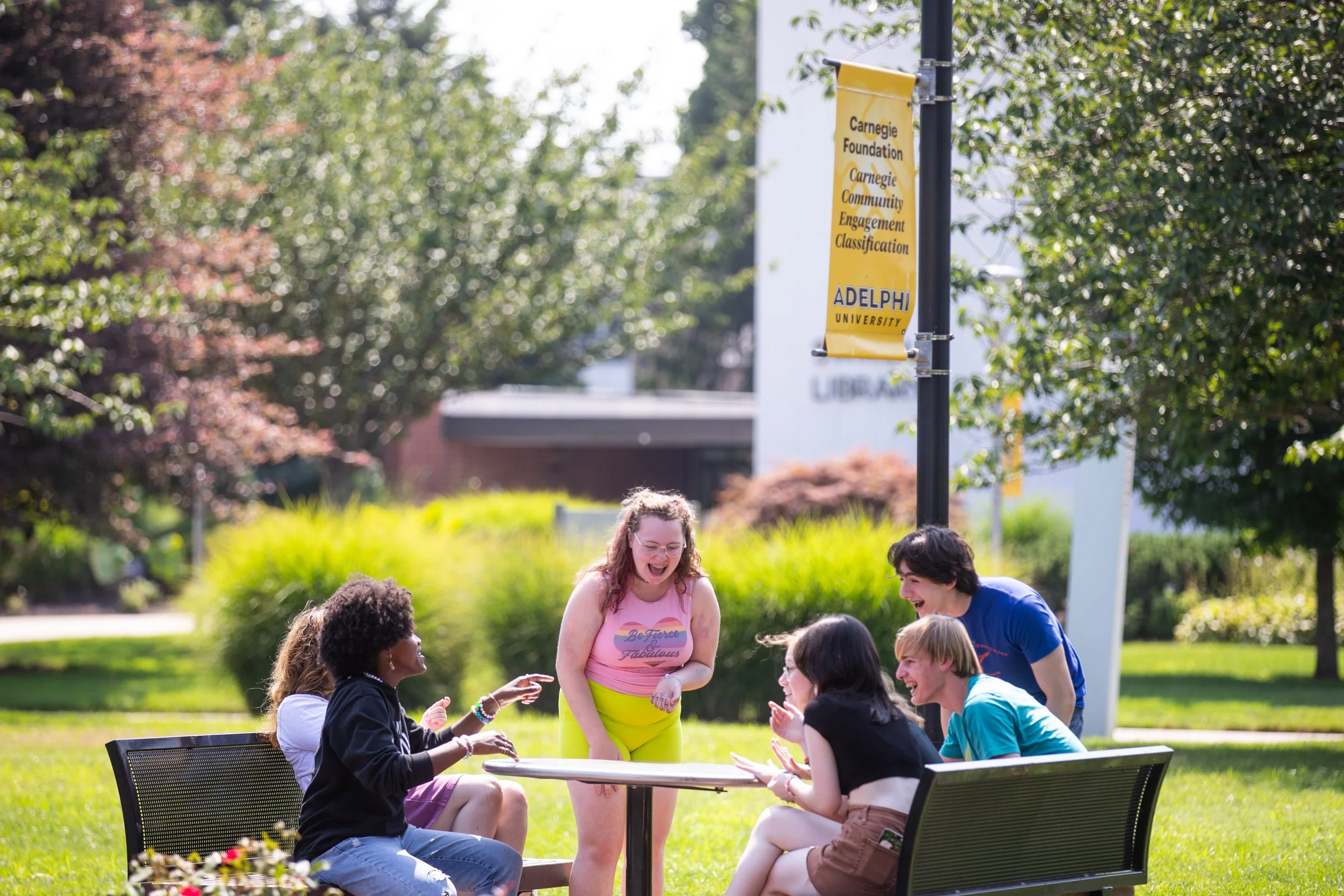 Group of diverse young people laughing and talking around a table outdoors on a sunny day at Carnegie Mellon University, with a yellow banner and trees in the background.