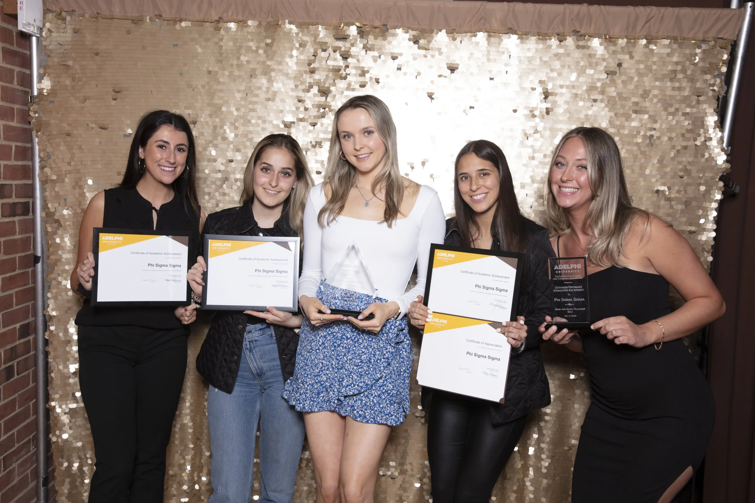 Group of five women standing in front of a gold sequin backdrop, holding awards and certificates, smiling for the camera.