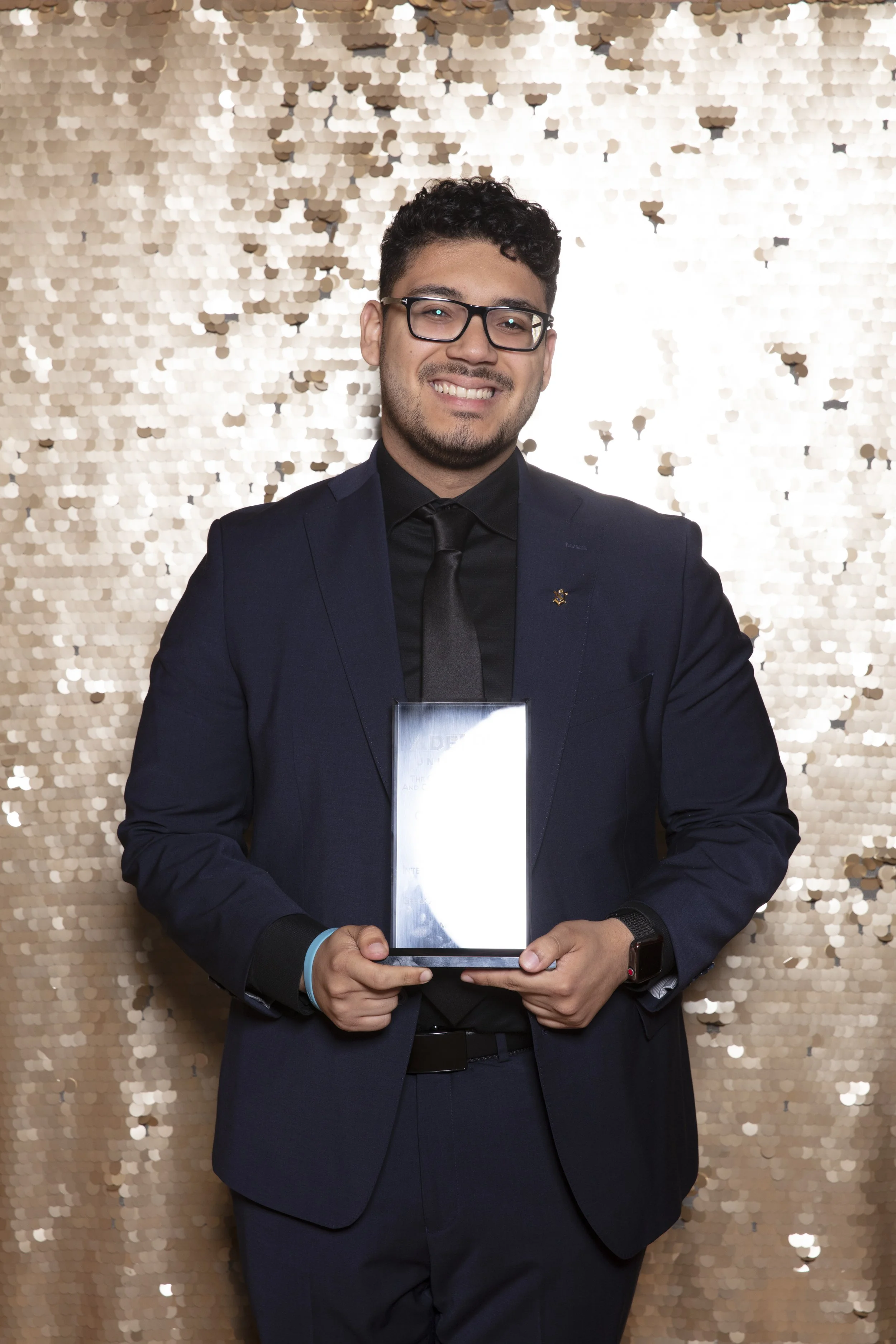A young man in a dark suit and tie, wearing glasses, smiling and holding a glass award, standing in front of a gold sequin backdrop.