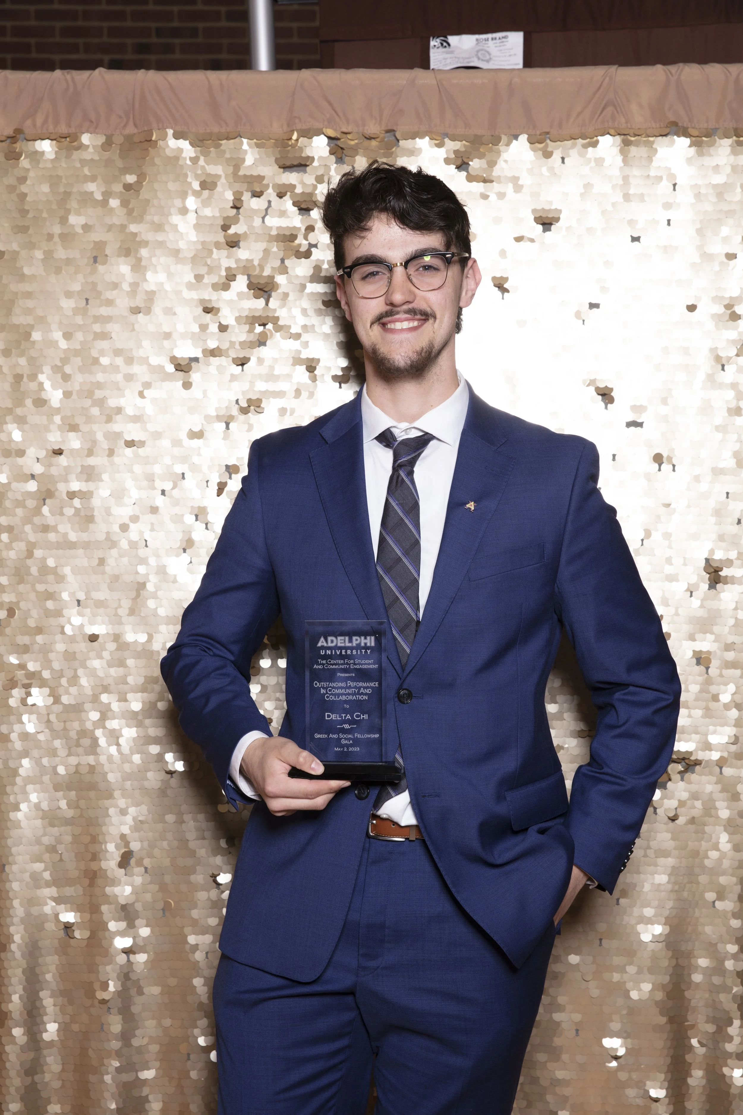Young man in a navy suit and glasses holding an award trophy, standing in front of a shiny gold sequin backdrop.